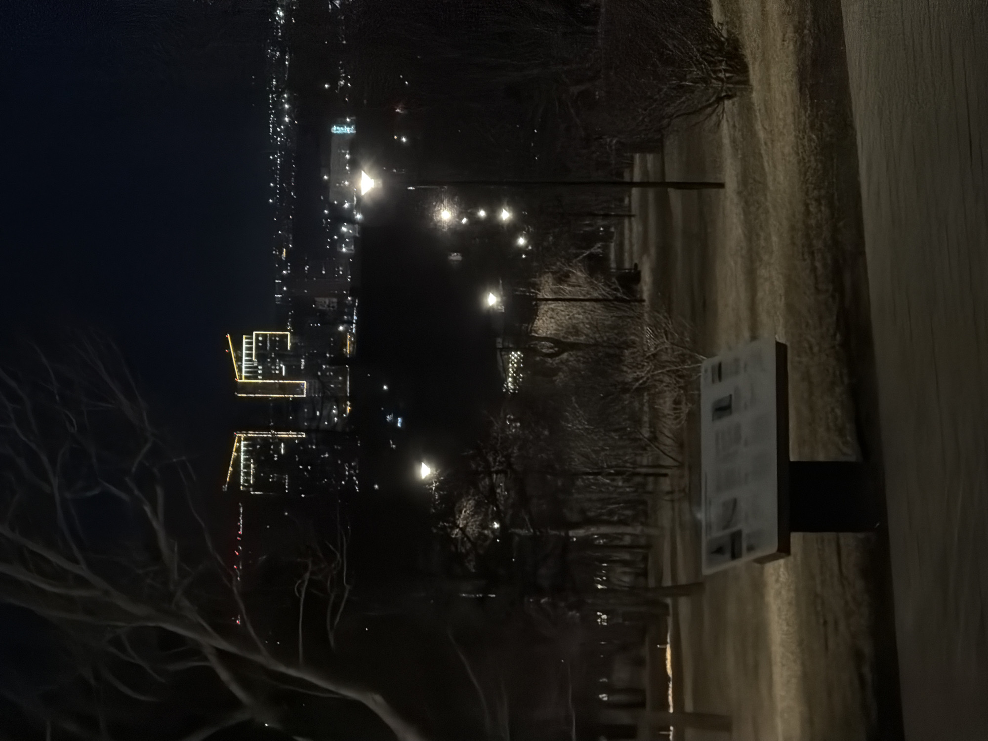 The image depicts a nighttime scene in Krasnoyarsk, Russia. The cityscape is visible in the background, illuminated with various lights including distinctive illuminated buildings. In the foreground, streetlights line a pathway. A sign is also present, mounted on a metal post. The surrounding environment appears to be a park or similar green space. The ground is covered with snow. Bare tree branches are visible.