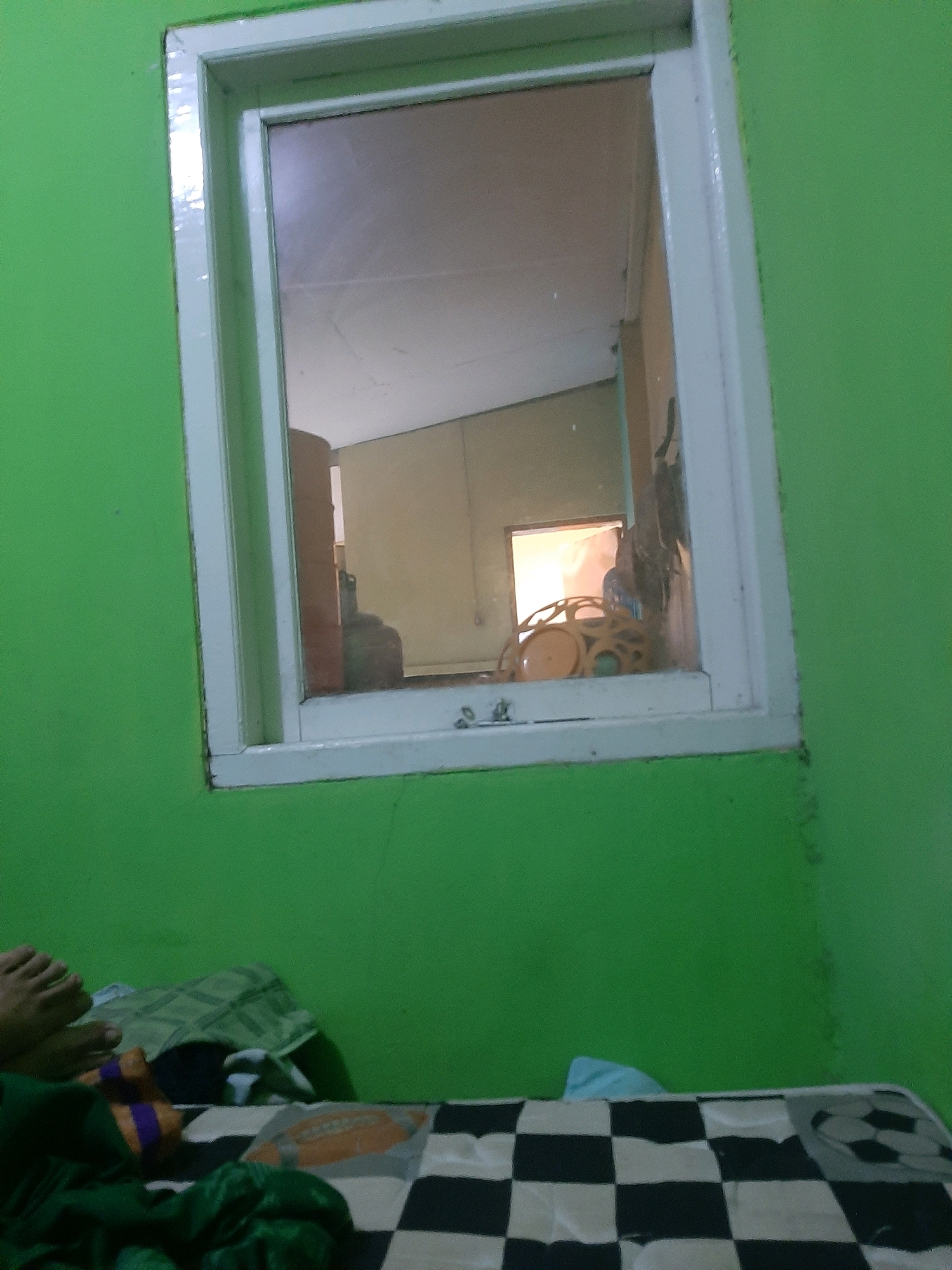 The image depicts an interior scene with bright green walls. A white-framed window occupies a significant portion of the frame, reflecting the interior of the room. The reflection shows what appears to be a ceiling and a doorway. Inside the window frame, on the windowsill, a tan-colored object with circular cutouts is visible, along with a dark, blurry object that could be a piece of furniture or a shadow. In the lower left portion of the image, a person's feet are visible, resting on a checkered surface. There is also green fabric draped over the checkered surface. The overall impression is of a modest, likely domestic, interior space. The specific location within Sumampir, Indonesia, cannot be determined from the visual information alone.