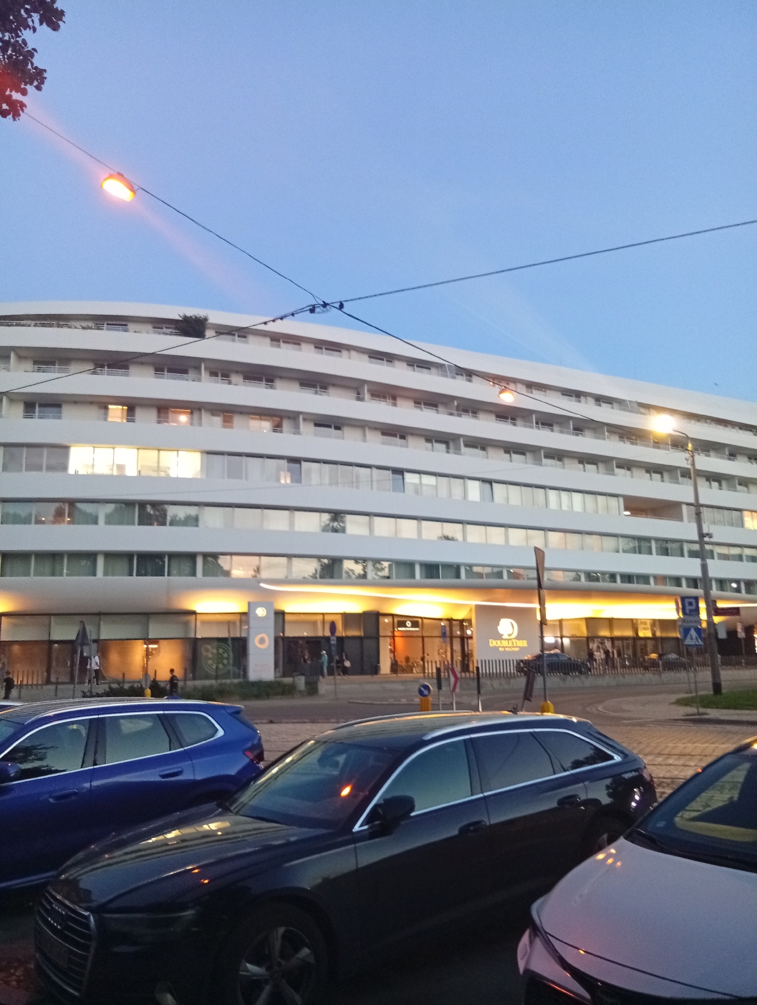 The image captures an exterior view of the DoubleTree by Hilton Wrocław hotel in Wrocław, Poland, at dusk. The modern, white facade of the multi-story hotel is illuminated by warm interior lights from its numerous windows and a bright, orange-hued awning over the ground floor entrance. A prominent DoubleTree by Hilton logo is visible on a white sign at the entrance. The hotel is situated next to a street where several cars are parked. In the foreground, two cars are parked closely together: a dark-colored Audi in the center and a lighter-colored vehicle to its right. A blue station wagon is parked to the left of the Audi. Streetlights are visible, one casting a bright glow from the right side of the frame and another, an orange streetlamp, is suspended from a wire in the upper left portion of the image. The sky is a clear, dusky blue with subtle cloud streaks. Power lines and street lamps are prominent features in the sky above the hotel. Pedestrians can be seen in the distance near the hotel entrance.