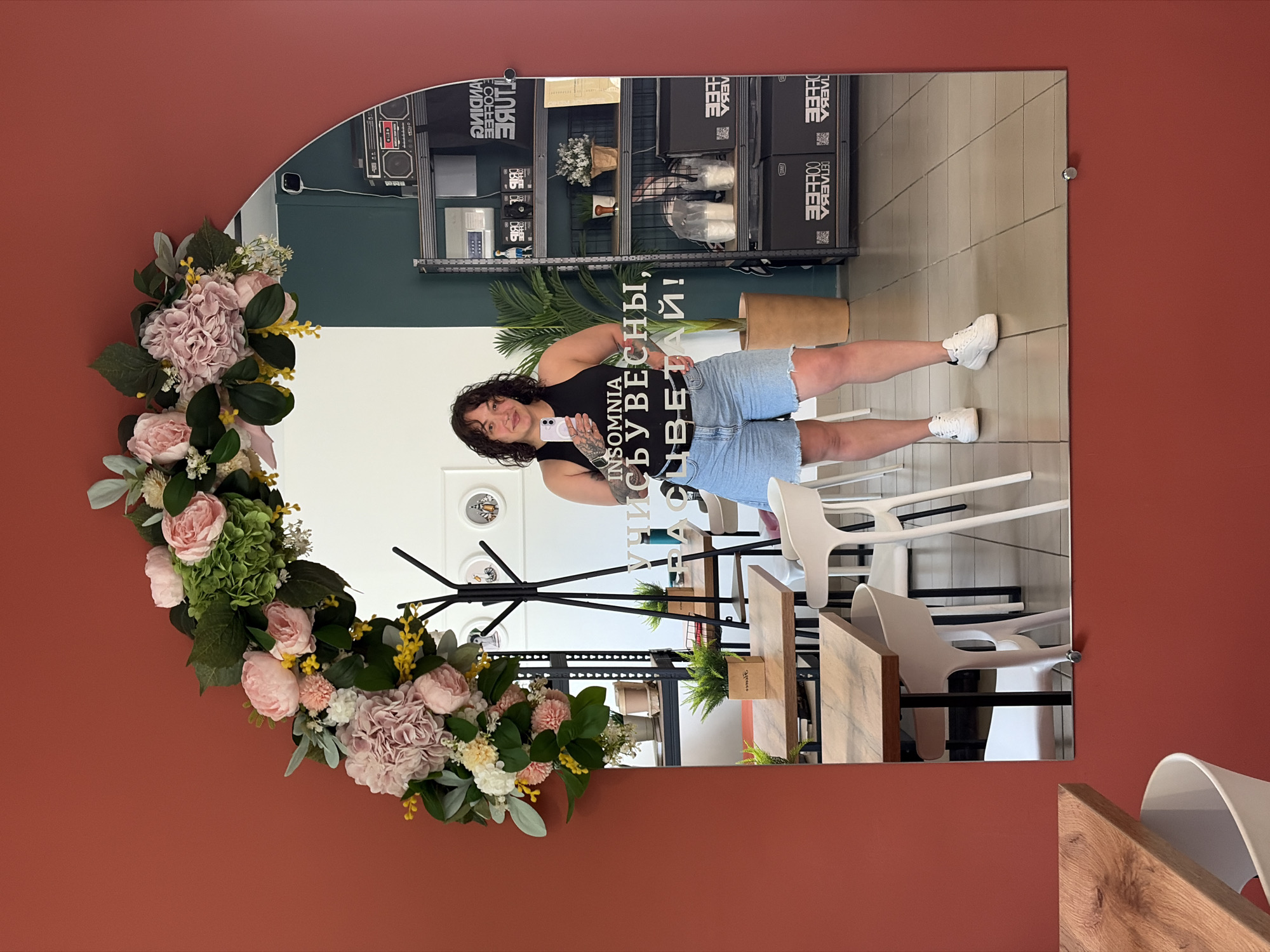 A woman stands in front of an arched mirror decorated with a floral garland. She is wearing a black tank top and denim shorts. The mirror reflects a cafe interior with tables, chairs, and shelving units. Text on the mirror, visible in reverse, reads "INSOMNIA. СУБЕСНЫ, ЦВЕТАЙ!". The cafe appears to be located in Chaikovsky, Russia, as indicated by the presence of the text in Russian and the general appearance of the establishment.