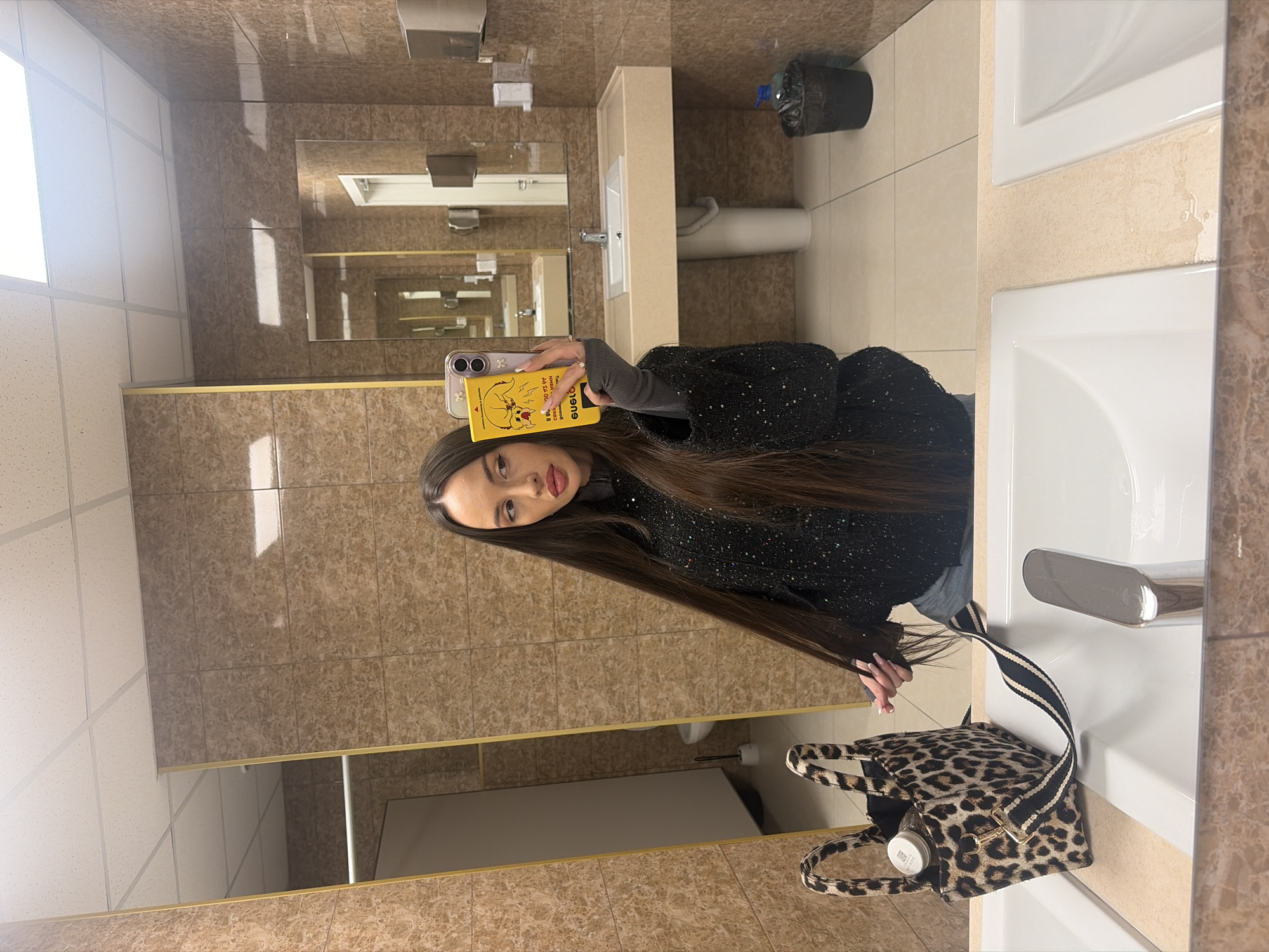 A woman is taking a selfie in a bathroom. She is holding a yellow smartphone with a cartoon design on the back. The bathroom has beige tile walls and white sinks. A leopard-print purse is on the sink next to her. A toilet and waste bin can be seen in the background, as well as a mirror. The location is difficult to ascertain with the available information, but it appears to be a public bathroom. There is no indication of Saint Petersburg, Russia.