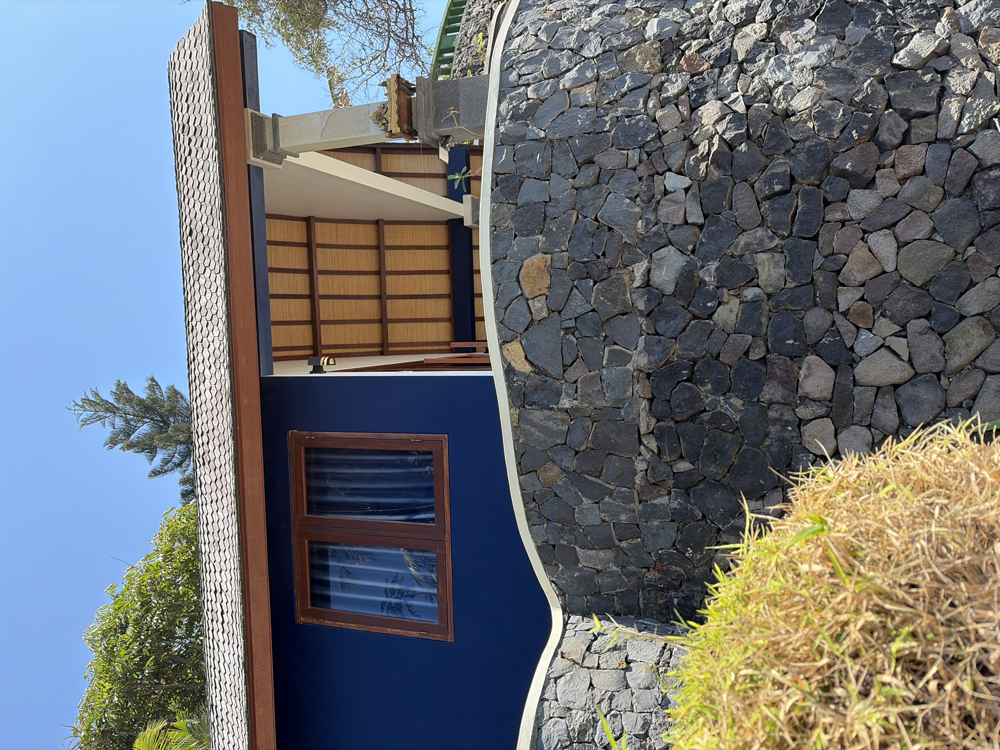 This is an exterior, eye-level, medium shot of a building in Abang, Indonesia, on a clear, sunny day. The building features dark blue walls, a wooden-framed window, and a tiled roof. A curved stone wall, constructed from irregularly shaped gray stones, is in the foreground, partially obscuring a clump of dry, yellowed grass. Part of a covered patio with bamboo ceiling panels is visible. The scene is devoid of any people or animals.