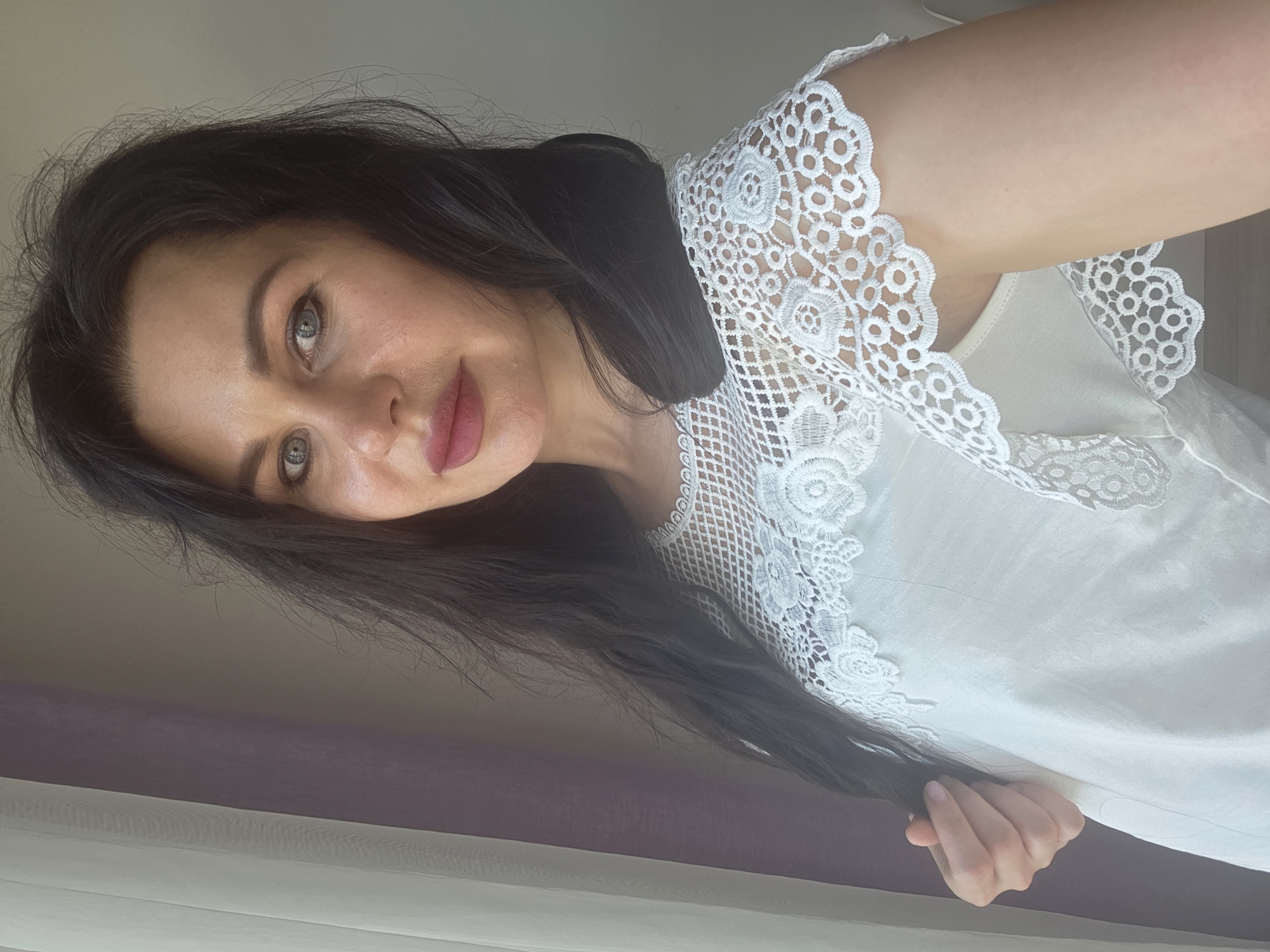 A close-up portrait of a woman with long, dark hair, fair skin, and light grey or blue eyes is displayed in a landscape orientation, appearing as if the subject is looking upwards and slightly to the left. She has subtle makeup, including pinkish-red lipstick. She is wearing a light-colored (white or off-white) top adorned with intricate white lace detailing along the right shoulder and neckline. Her right hand is visible in the lower right portion of the frame, gently holding a strand of her dark hair. The background reveals an indoor setting with natural light, suggesting it is daytime. On the right side, a sheer white curtain is visible, layered over a darker, possibly purple or plum-colored curtain or wall. The woman maintains a calm and serene expression, with a gentle gaze directed towards the viewer. The image was taken in Sochi, Russia. There is no visible text.