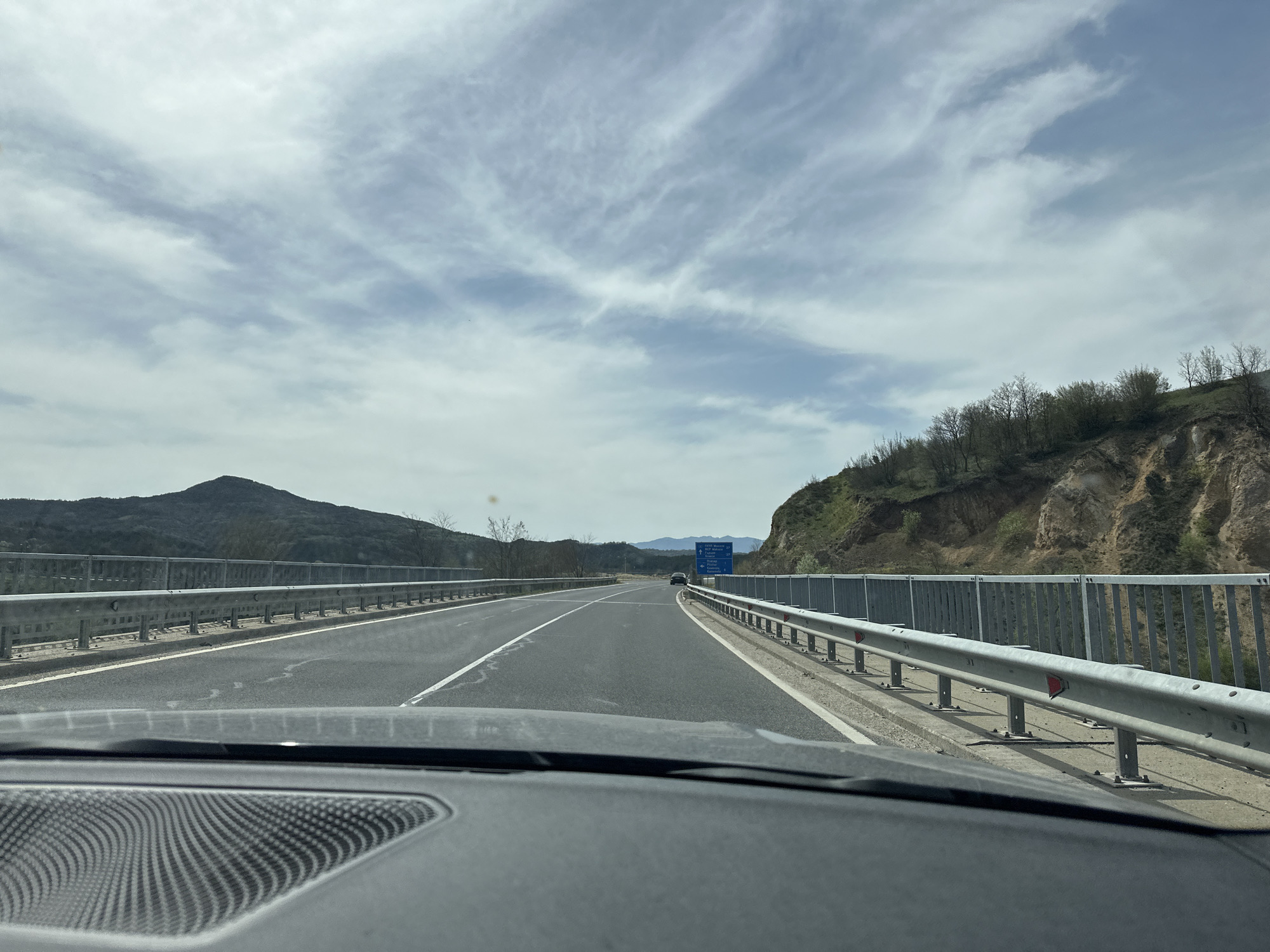 Captured from within a vehicle, the image presents a view of a highway cutting through a hilly landscape. The road is characterized by two lanes running in each direction, separated by a dashed white line, and bordered by guardrails on either side. Off in the distance is a highway sign of Zagorsko, Bulgaria. Mountains are visible in the background. The sky is cloudy. The vehicle's dashboard is visible in the foreground.