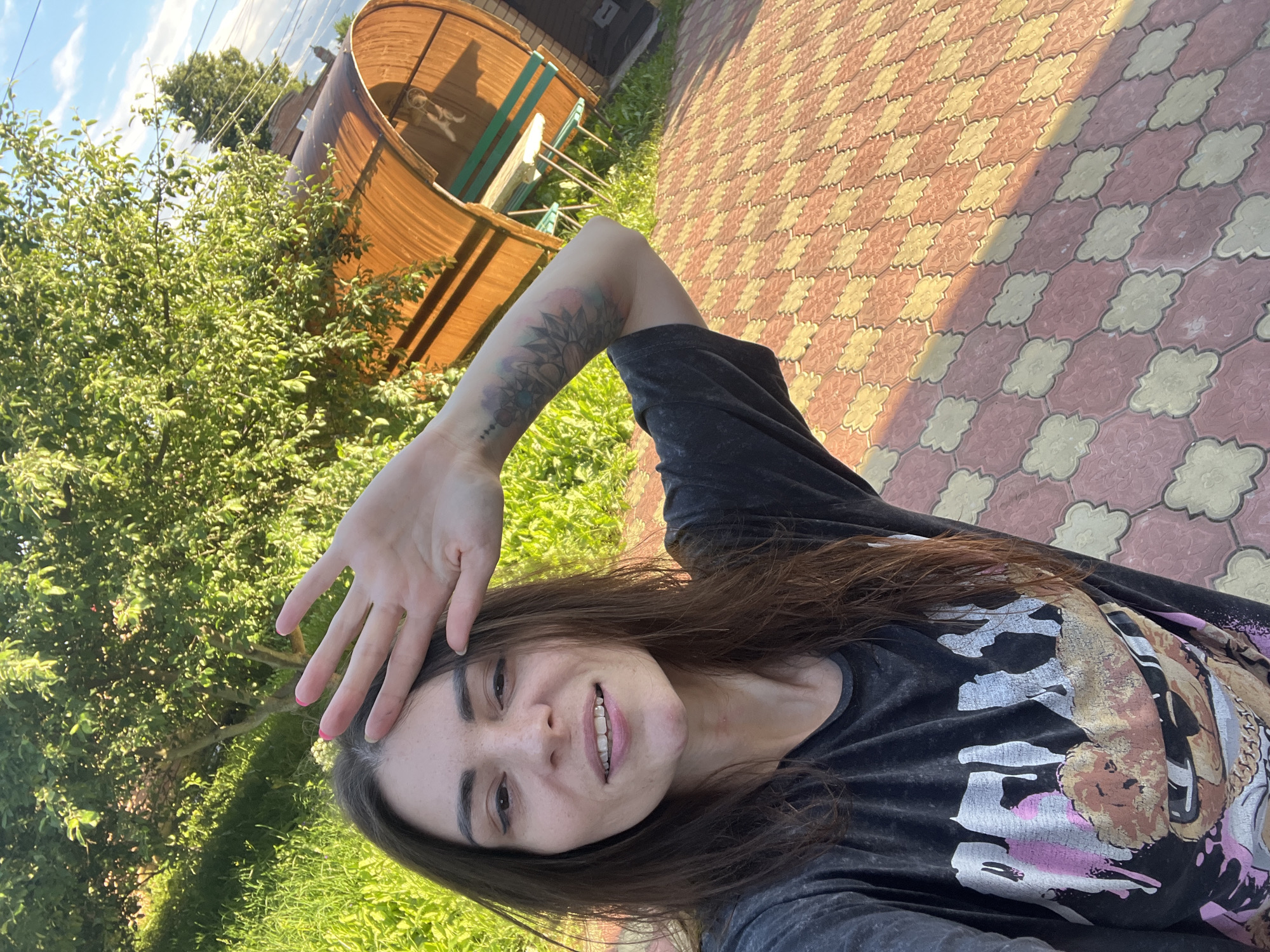 The image shows a young woman taking a selfie outdoors. She is wearing a dark t-shirt with a graphic print, and her right arm is raised to shield her face from the sun. She has long, dark brown hair and is smiling. Tattoos are visible on her arm.
The setting appears to be a backyard or garden area. To the left of the woman is lush green foliage and trees. Behind her and to the right, there is a cobblestone pathway made of red and yellow patterned pavers. A curved wooden structure, possibly a gazebo or seating area, is visible in the background, with green benches inside.
The lighting suggests it is daytime, likely late afternoon given the angle of the shadows cast on the pathway. The sky is blue with some clouds. The overall mood of the image is casual and relaxed, with the woman appearing happy and enjoying the sunny weather. No visible text is present in the image. The location context of Krasnoe-na-Volge, Russia, suggests a rural or semi-rural setting.