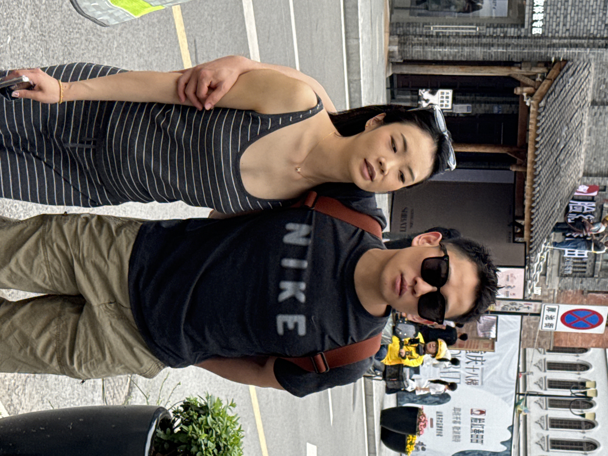 A medium shot shows a man and a woman. The man is wearing a black t-shirt with “NIKE” printed on it in white, brown pants, a brown backpack, and black sunglasses. The woman is wearing a striped tank top. They stand on a paved street next to a planter, in front of buildings and banners of shops. The sign “厚池街” is seen on a building in Yuzhong District, China.