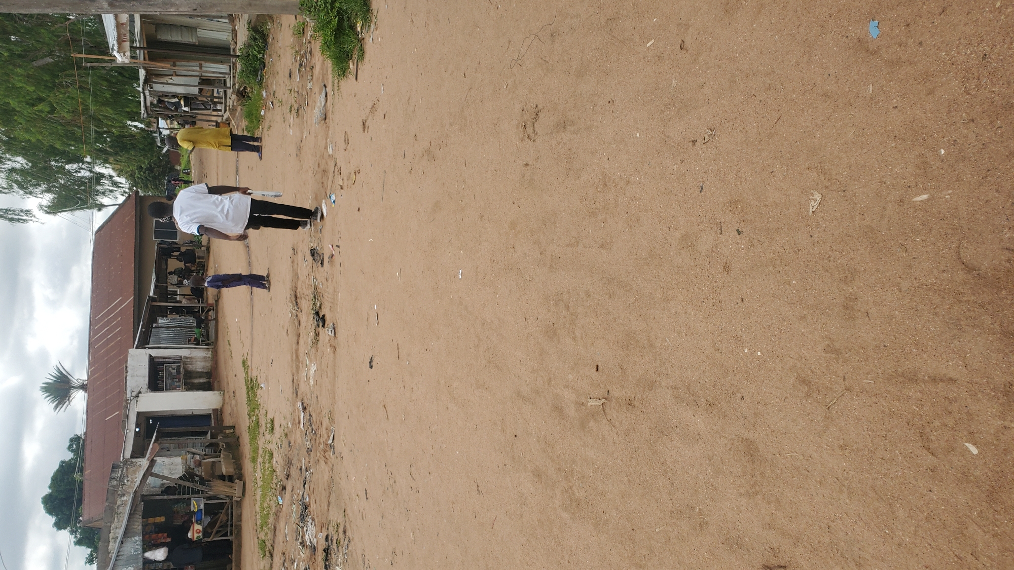 In Jalingo, Nigeria, a dusty, unpaved road is the setting for a scene of daily life. The road is lined with informal shops and structures, some with corrugated metal roofs and others with more rudimentary coverings. Debris, including plastic bags and other trash, is scattered across the sandy ground, indicating a less-than-pristine environment.
Several individuals are present. In the foreground, a young man wearing a white t-shirt and dark pants walks away from the camera, carrying a white object in his hand. A young child in a dark blue outfit stands a few feet in front of him, facing the shops. Further down the road, a person in a yellow garment is walking towards the shops. Other individuals appear to be inside or near the shops, some engaged in activities not clearly discernible from this angle. The background features a few trees and a cloudy sky, typical of a warm climate. The overall impression is one of a bustling, yet somewhat unkempt, local market area.