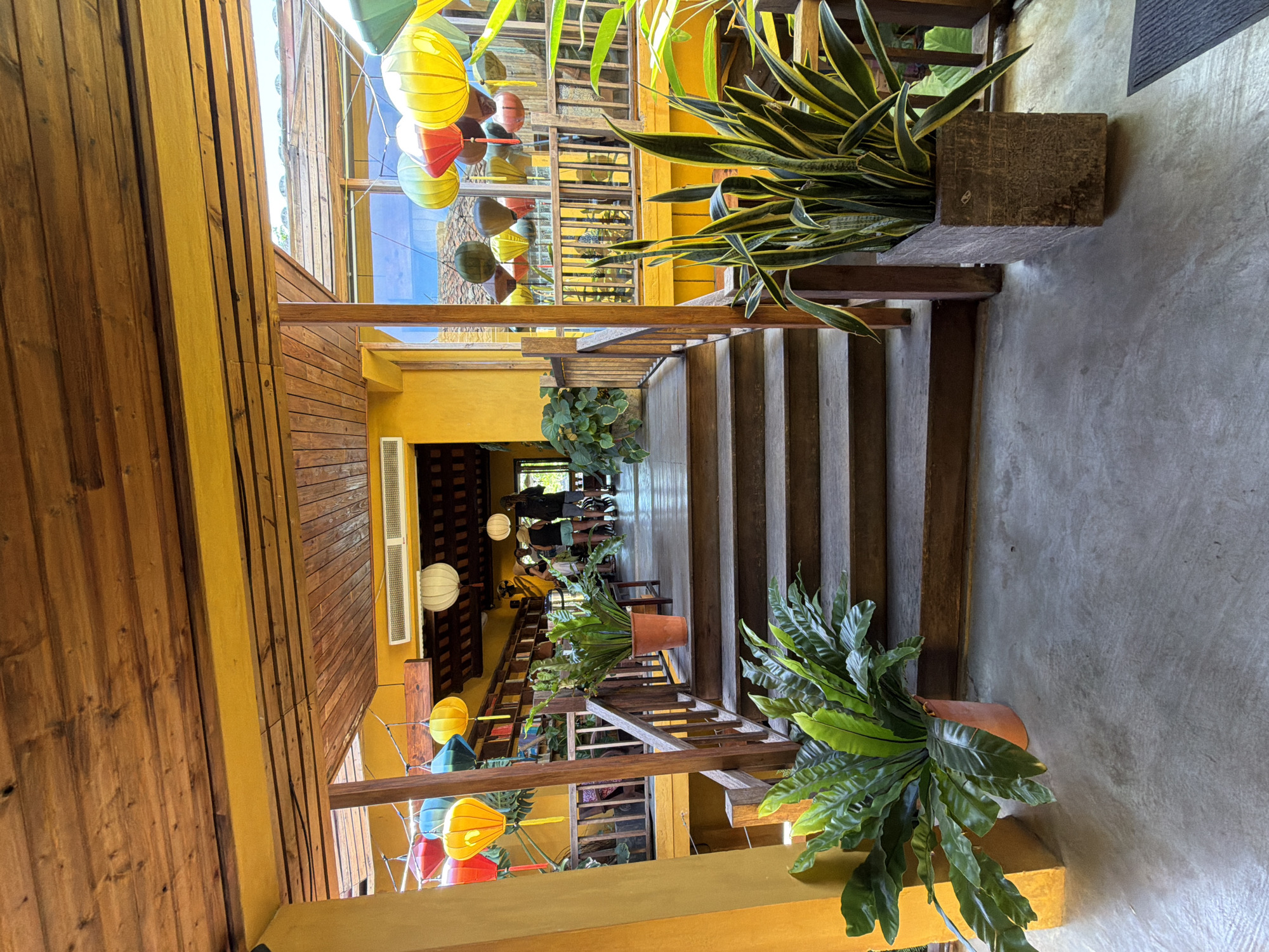 The image depicts a view of a wooden staircase and walkway within a building in Hội An Ward, Vietnam. The architecture features prominent yellow walls and a wooden ceiling with exposed beams. Decorative lanterns in various colors, including red, yellow, blue, and green, are suspended from the ceiling and hang throughout the space. Potted plants, including a bird's nest fern and a snake plant, are placed along the walkway and near the stairs. In the background, several individuals are visible standing on a higher level, some interacting and others appearing to observe the surroundings. The scene suggests a relaxed, perhaps commercial or hospitality-oriented setting, with the unique lanterns indicating a Vietnamese cultural aesthetic.