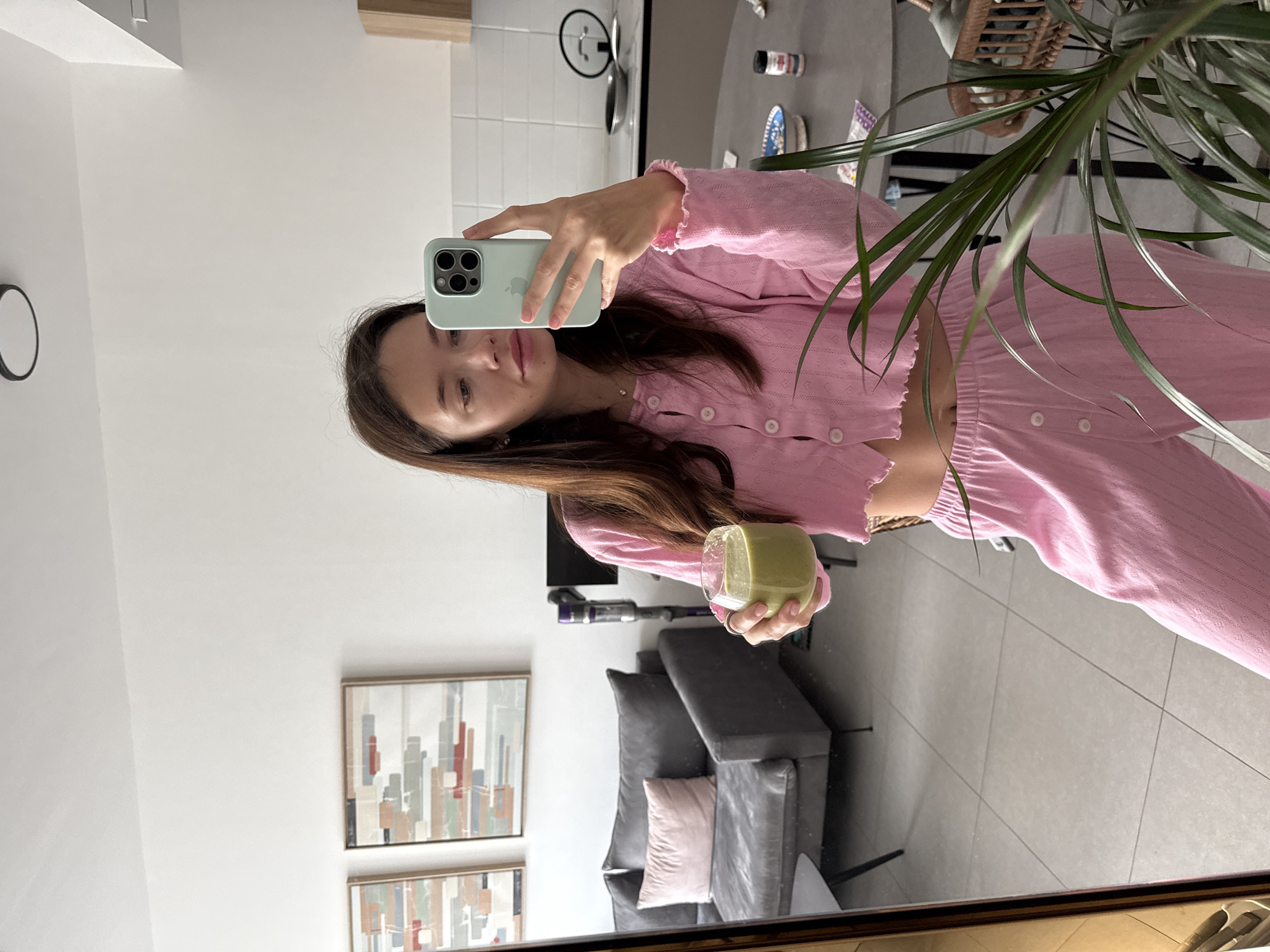 A young woman is captured in a mirror selfie, wearing a light pink, two-piece loungewear set consisting of a cropped, long-sleeved button-up top with ruffled cuffs and matching pants. She holds a light green iPhone with three camera lenses in her right hand, angled to capture her reflection. In her left hand, she holds a clear glass containing a green beverage, possibly a smoothie or matcha. Her long, dark brown hair cascades over her shoulders, and she has a calm expression with a slight smile. The visible Apple logo on her phone case is the only discernible text.
The reflection reveals a modern and tidy indoor living space, likely an apartment in Ramat Gan, Israel. To the woman's right, a tall potted plant with long green leaves partially obscures the view. The background features a grey dining table with a small black clock, spice jars, and a decorative patterned plate. Further into the room, a grey sofa with a light pink throw pillow is visible. On the white wall above the sofa, two framed abstract artworks with geometric shapes in muted colors (grey, red, beige) are displayed. A Dyson stick vacuum cleaner stands against the wall. The floor is covered with light grey tiles. The upper left corner of the reflection shows a white tiled wall, hinting at a nearby kitchen area. The scene depicts a casual, relaxed moment at home, likely during the daytime.
