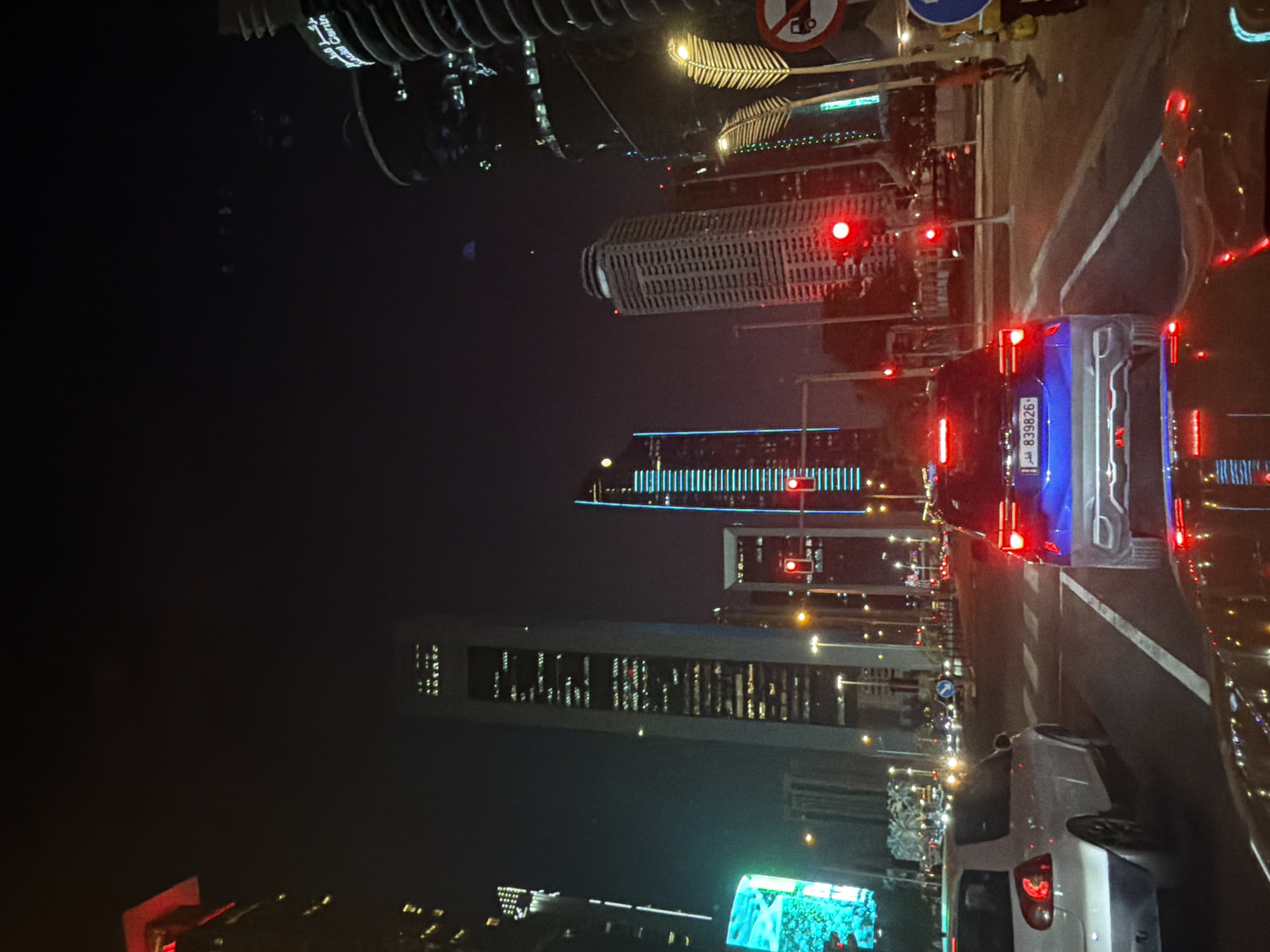 This forensic description details an urban night scene from the perspective of a vehicle stopped at a red traffic light. The setting is a city in Qatar, specifically indicated by the Arabic text "قطر" on the license plate of the blue SUV directly ahead. Given the prominent modern skyscrapers, the location is likely Doha's West Bay district.
In the immediate foreground, reflections of the dashboard and internal vehicle lights are visible on the windshield, including indistinct red glows. A white two-door hatchback, possibly a Volkswagen Golf, is positioned to the left, slightly ahead, with its red taillight visible. Directly in front, a blue compact SUV (possibly a Volkswagen T-Roc) is stopped. Its rear license plate, "قطر 839826", is clearly legible, confirming the country. The SUV's brake lights are illuminated red.
The road features multiple lanes demarcated by white solid and dashed lines, along with a marked pedestrian crossing. Three prominent red traffic signals are visible, two above the intersection ahead and one on a pole to the right, indicating the stop. Numerous streetlights cast a yellow-orange glow on the road.
On the right side of the road, unique decorative streetlights shaped like stylized yellow leaves with internal illumination are present. Several road signs are also visible: a circular red sign with a black truck (indicating "No Trucks") and a circular blue sign with a white arrow (likely a directional or mandatory instruction sign). A person wearing a yellow or light-colored vest is partially visible on the far right pavement, near the "No Trucks" sign.
The background is dominated by a cluster of towering, illuminated skyscrapers against a dark night sky. A tall, slender, light-grey building with multiple illuminated windows stands centrally. To its right, another skyscraper features distinctive vertical blue strip lighting. Further to the right, a building with a textured, possibly spiraling or ridged facade displays partial Arabic and English text, including "Financial Centre," suggesting a commercial district. Other illuminated buildings and distant city lights fill the horizon. The overall scene depicts a busy, modern, and well-lit metropolitan area at night.