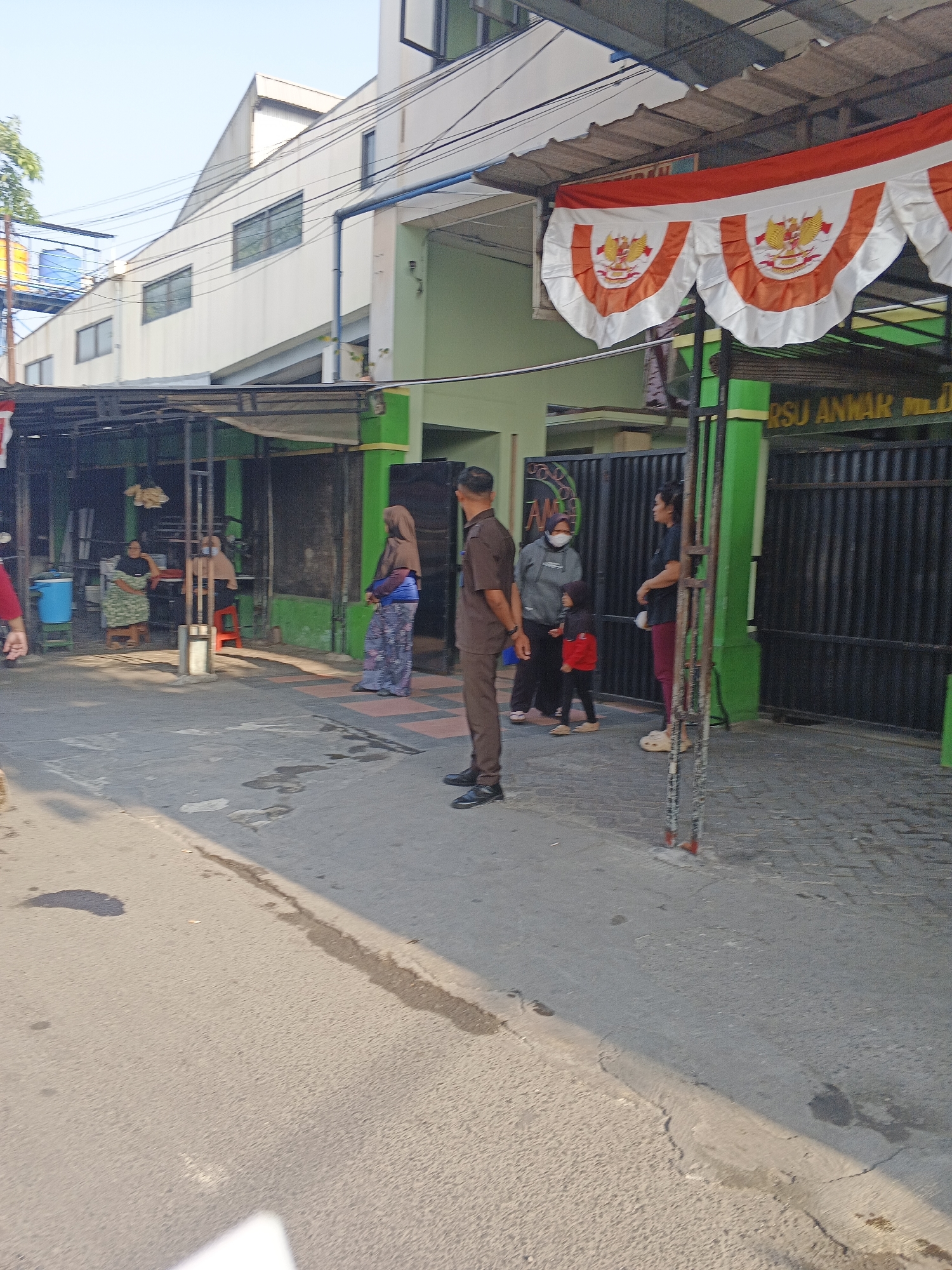 The image depicts an outdoor scene in front of a building that appears to be a hospital, with a sign that reads "RSU ANNAR". The building has a light green exterior and is partially shaded by a canopy adorned with red and white pennant banners featuring the Garuda Pancasila emblem. In the foreground, a paved road with some water stains and shadows is visible.
Several individuals are present. On the left, near the entrance, two women are seated at tables, with a blue cooler box nearby. A man in a brown uniform stands near the center of the frame, facing towards the right. To his right, a woman wearing a hijab and a patterned skirt is standing. Further to the right, a group consists of a woman in a gray hoodie and mask, a young child in a red jacket, and another woman in maroon pants. A black metal fence with a green post separates the paved area from the building. The sky above is clear and blue. The overall atmosphere suggests a public area, possibly a waiting or service area outside the RSU Annar in Krian, Indonesia.
