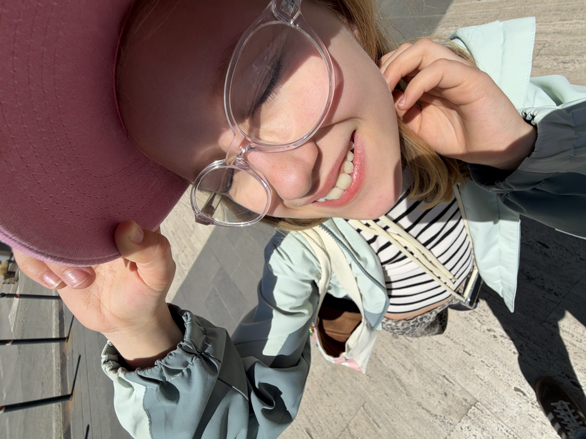 A photograph, taken from above, shows a fair-skinned woman smiling with her eyes partially closed. She is wearing clear-framed glasses and a pink hat. She has fair hair. She is wearing a striped shirt under a jacket. The jacket is light blue with grey sleeves. She is holding the edge of the pink hat. The background shows a beige-colored cobblestone surface. In the bottom right corner, a single foot is visible in a brown shoe. The location appears to be San Gimignano, Italy, given the stone paving material. The photograph appears to have been taken in daylight.