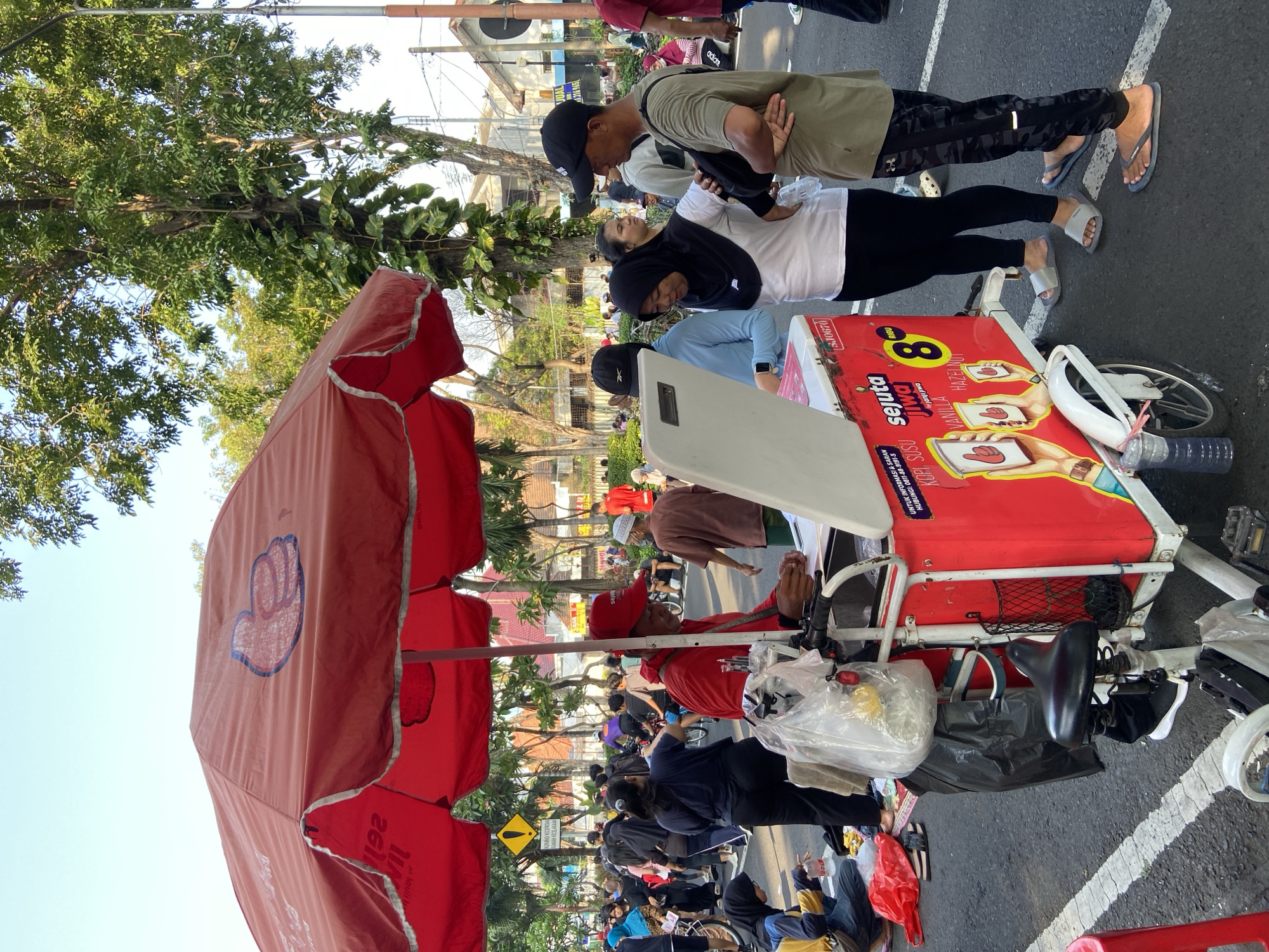 The image captures a bustling street scene in Surabaya, Indonesia, centered around a food vendor. The vendor, wearing a red shirt and cap, operates a red cart displaying advertisements for 'Janji Jiwa' coffee, priced at 8,000 Indonesian Rupiah (IDR). A red umbrella with a logo provides shade.
Several people are gathered around the cart, appearing to be customers. They include a man in an olive green shirt and black patterned pants, a woman in a white shirt and black pants, and another person in a light blue shirt. Many people are sitting on the ground.
The scene takes place on a paved street, likely in a business or public area, with white painted parking space lines marking parking spaces or driving lines. Buildings and trees are visible in the background.
