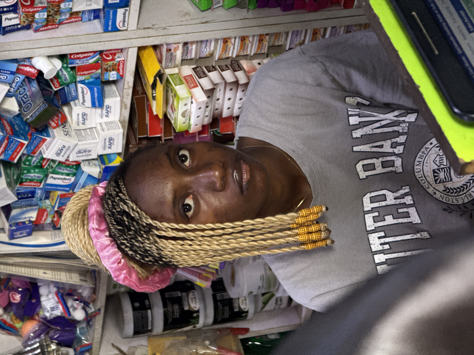 The image depicts a woman with braided hair, wearing a gray t-shirt with "COLUMBIA" and "CHARLESTON" printed on it, standing in what appears to be a small shop in Abuja, Nigeria. Shelves filled with various products, including toothpaste, breath mints, and other household items, surround her. She is looking directly at the camera. A tablet with a bright green case is partially visible in the upper right corner.