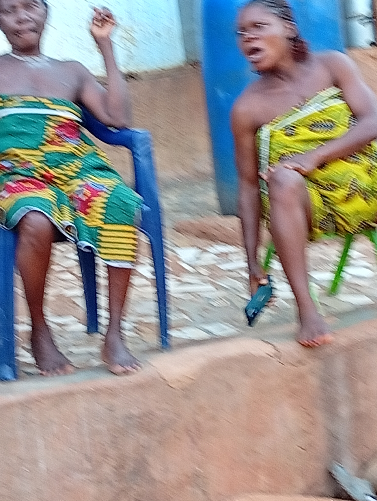 In Ogidi, Nigeria, two barefoot women sit outside. The woman on the left sits in a blue plastic chair, clad in a colorful green and red fabric wrap. She holds her right hand up. The woman on the right sits in a green plastic chair, wearing a yellow and green fabric wrap. She has her mouth open. A blue object resembling a phone lies on the ground near her feet. They sit on a raised earth platform near a blue plastic container.