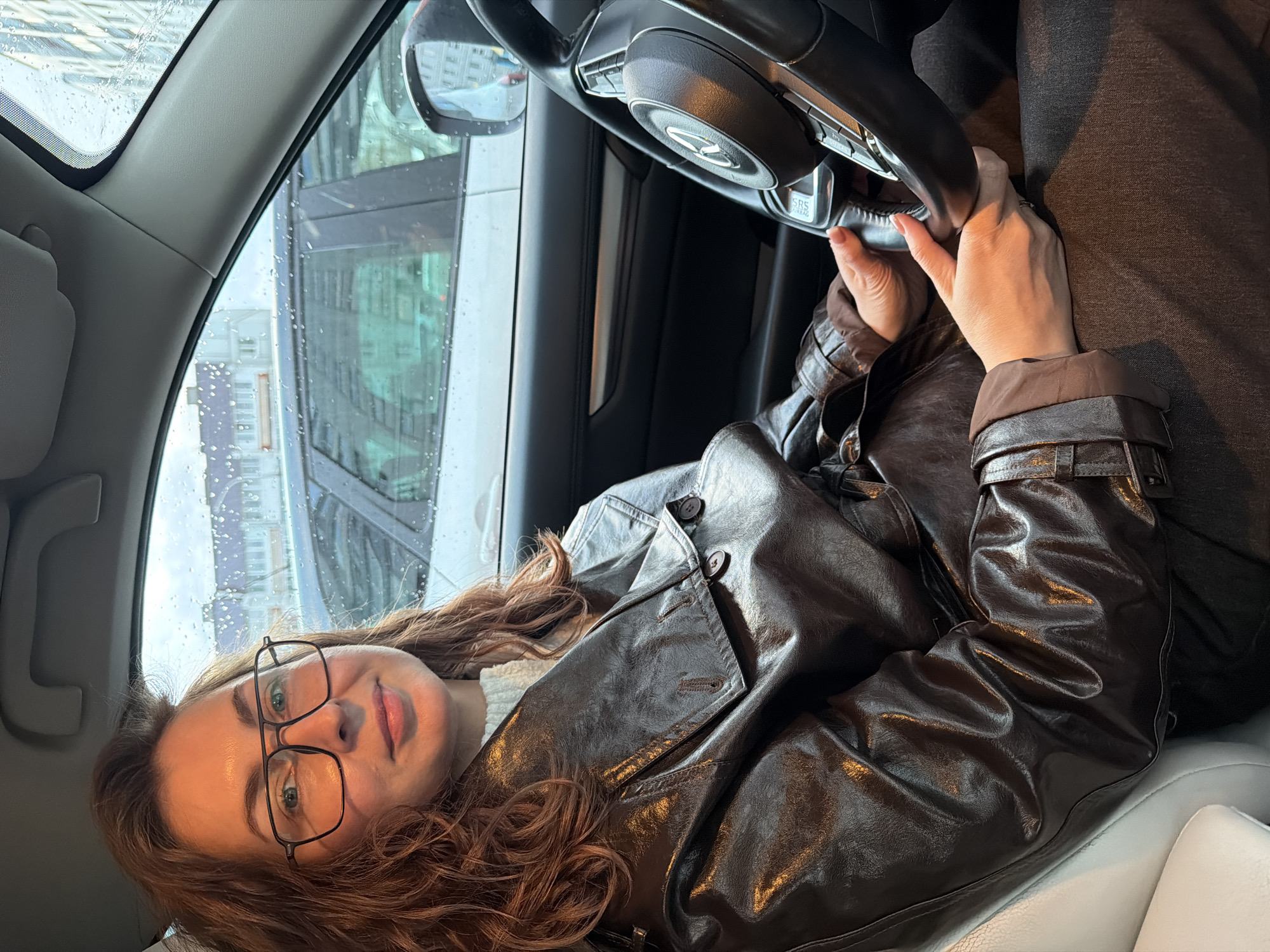The original image, oriented vertically, captures a woman in the driver's seat of a car, looking directly at the viewer with a slight smile. She has long, wavy, reddish-brown hair and wears dark-rimmed glasses. Her attire includes a glossy, dark brown leather-like trench coat over a lighter-colored top. Her hands are positioned on the steering wheel, which prominently displays a Mercedes-Benz logo and the text "SRS AIRBAG" near the controls.
The car's interior features a light-colored seat and dark dashboard elements. Through the front and side windows, raindrops are clearly visible, indicating rainy or recently rainy weather. In the background, out of focus, are multi-story urban buildings, suggesting a city environment. The lighting is soft daylight, highlighting the textures of her jacket and hair. The scene depicts a calm moment, likely a selfie or portrait taken while the car is stationary in Chaikovsky, Russia.
