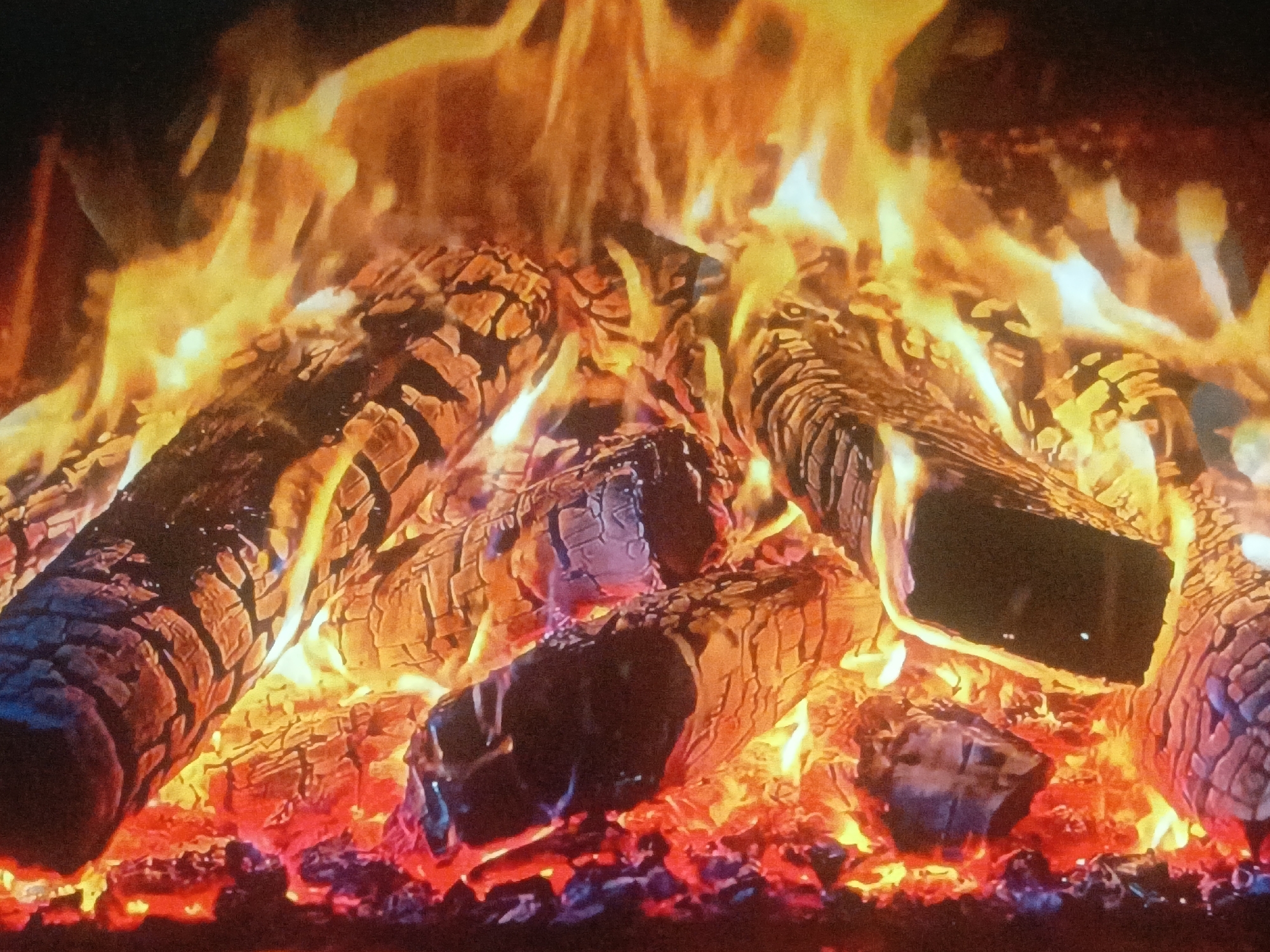 The image shows a close-up of a roaring fire with several logs burning intensely. The flames are predominantly yellow and orange, with some white flashes, extending upwards. The logs are dark brown with visible cracks and charred surfaces. The fire is emanating from a mass of embers and ash. No people are visible in the image. The location is not discernible from the visual information provided.