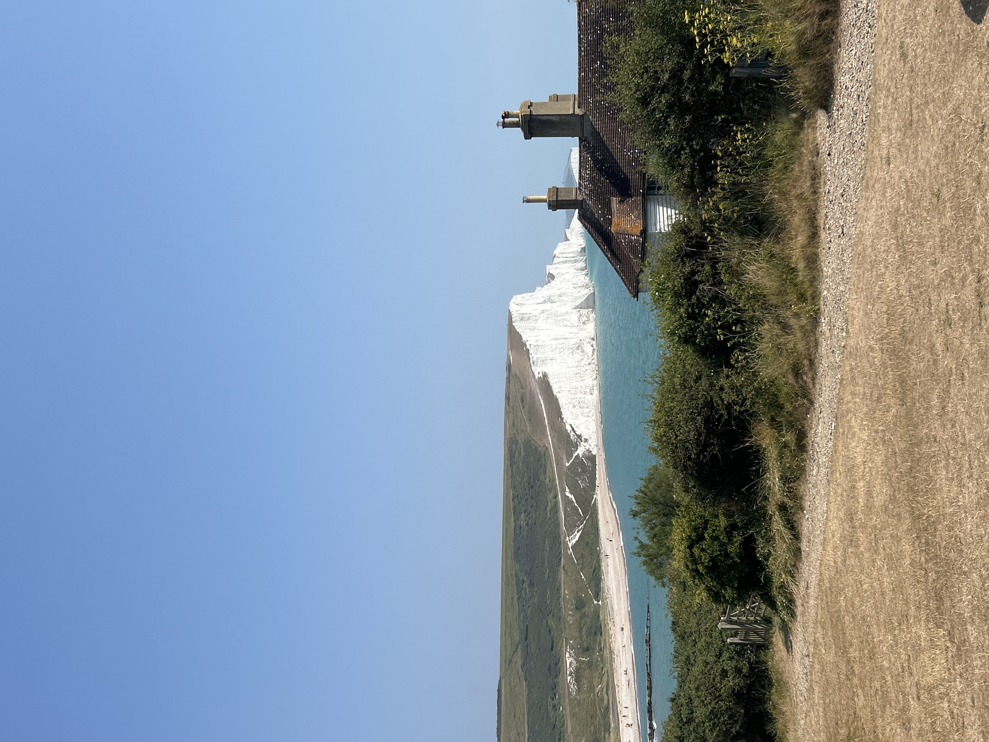 The image depicts a scenic coastal view of the Seven Sisters cliffs in Seaford, United Kingdom. In the foreground, dry, sun-bleached grass covers the ground, with a gravel path leading towards a wooden gate and dense green shrubbery. Partially visible behind the foliage is a white building with a brown tiled roof and two chimneys. The middle ground is dominated by the turquoise sea, which meets a pebble beach dotted with a few small figures. Beyond the beach, the iconic white chalk cliffs of the Seven Sisters stretch into the distance, meeting a clear blue sky. The overall impression is one of a bright, sunny day in a coastal landscape.