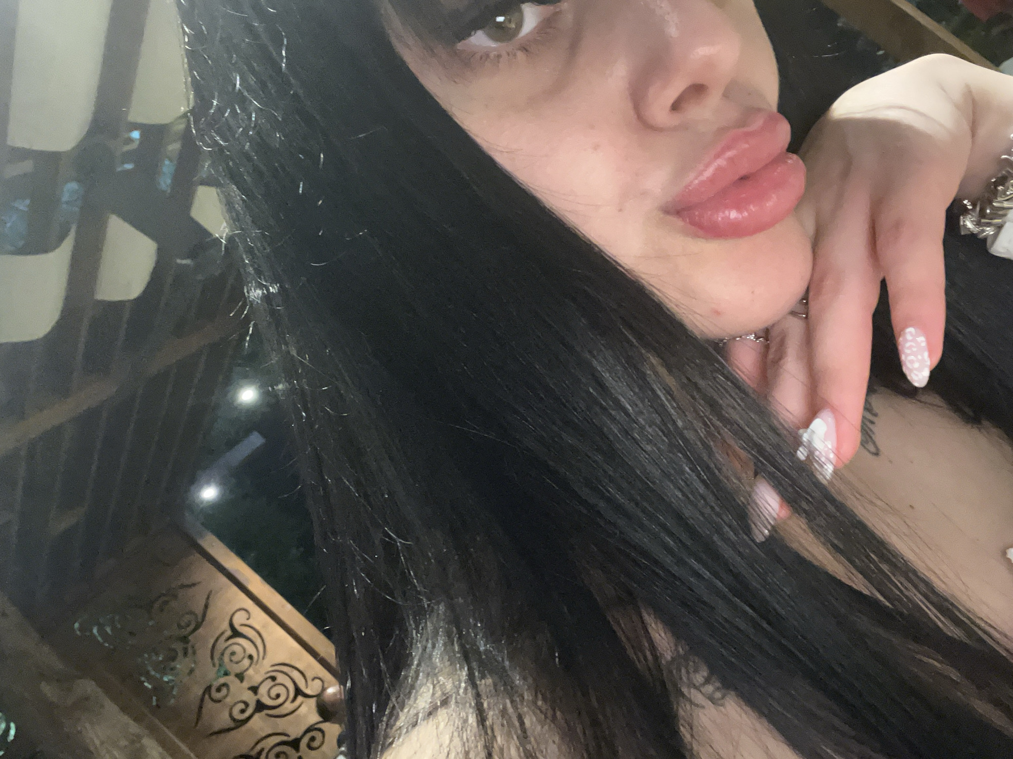 The image is a close-up selfie of a young woman with long, dark hair. She is wearing lip gloss and has a tattoo visible on her chest. Her nails are manicured with white and pink polish. She is wearing a silver ring on her finger. The background suggests she is outdoors, possibly under a wooden pergola or canopy, with lights visible in the distance. The overall lighting is dim. There are no discernible landmarks that would specifically place this image in Plovdiv, Bulgaria.
