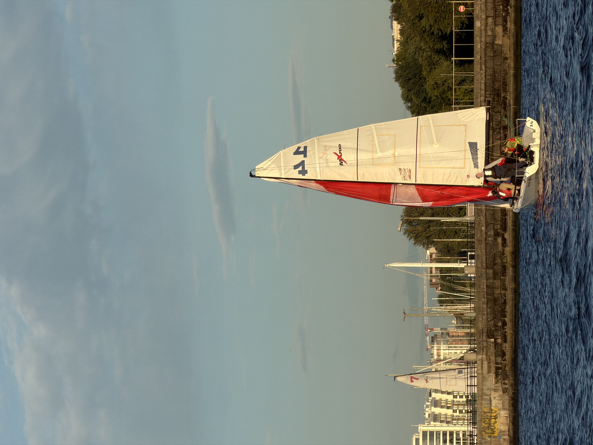 A sailboat with a white and red sail navigates on a blue body of water. The sail has the number "44" in dark blue and the "MAXI 700" logo in red. Four individuals are on board the sailboat, wearing life vests in various colors. In the background, a stone embankment runs horizontally, with buildings of various heights and architectural styles lining the shore. Trees and a fence are also visible behind the embankment. Further in the distance, masts of other boats and a crane are present, suggesting a harbor or marina. The sky is a light blue with scattered clouds. This scene is located in Saint Petersburg, Russia, likely along the Neva River or a connected waterway, given the urban development and maritime activity.