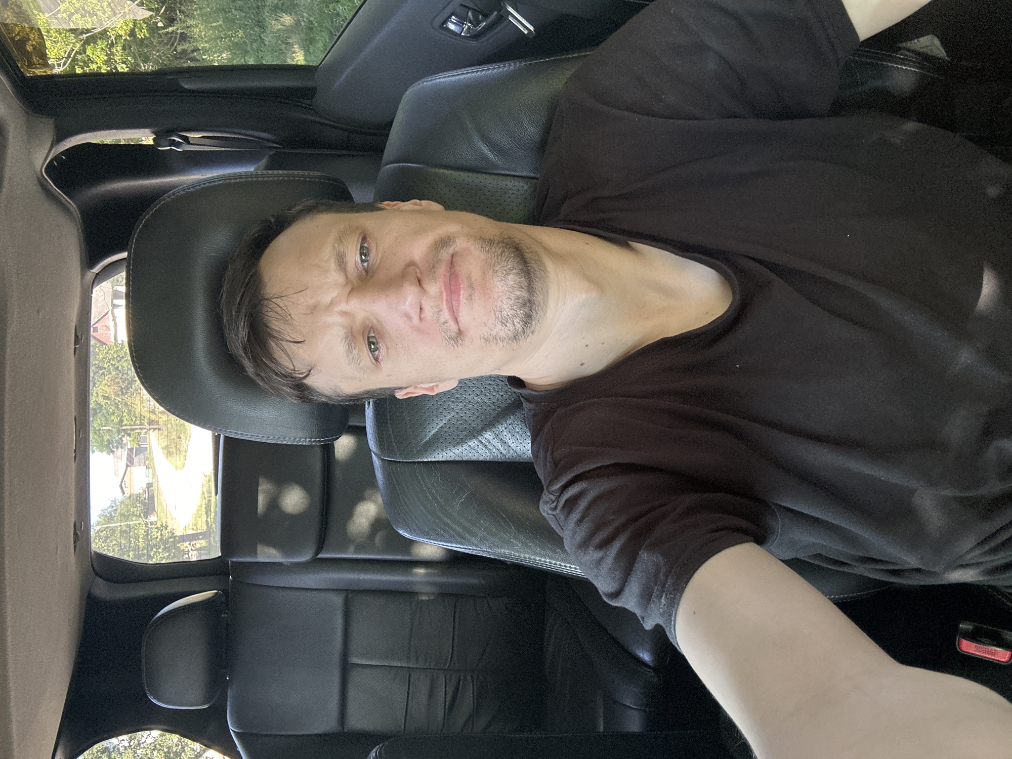 A man with short brown hair and light green eyes is sitting in the driver's seat of a vehicle. He has a stubble beard and is wearing a black t-shirt. His eyebrows are furrowed, and his mouth is slightly open, giving him a questioning or slightly displeased expression. The interior of the vehicle is visible, with black leather seats and headrests. Through the rear window, a dirt road curves past a couple of houses and trees, suggesting a rural or semi-rural setting. The overall lighting suggests it is daytime.