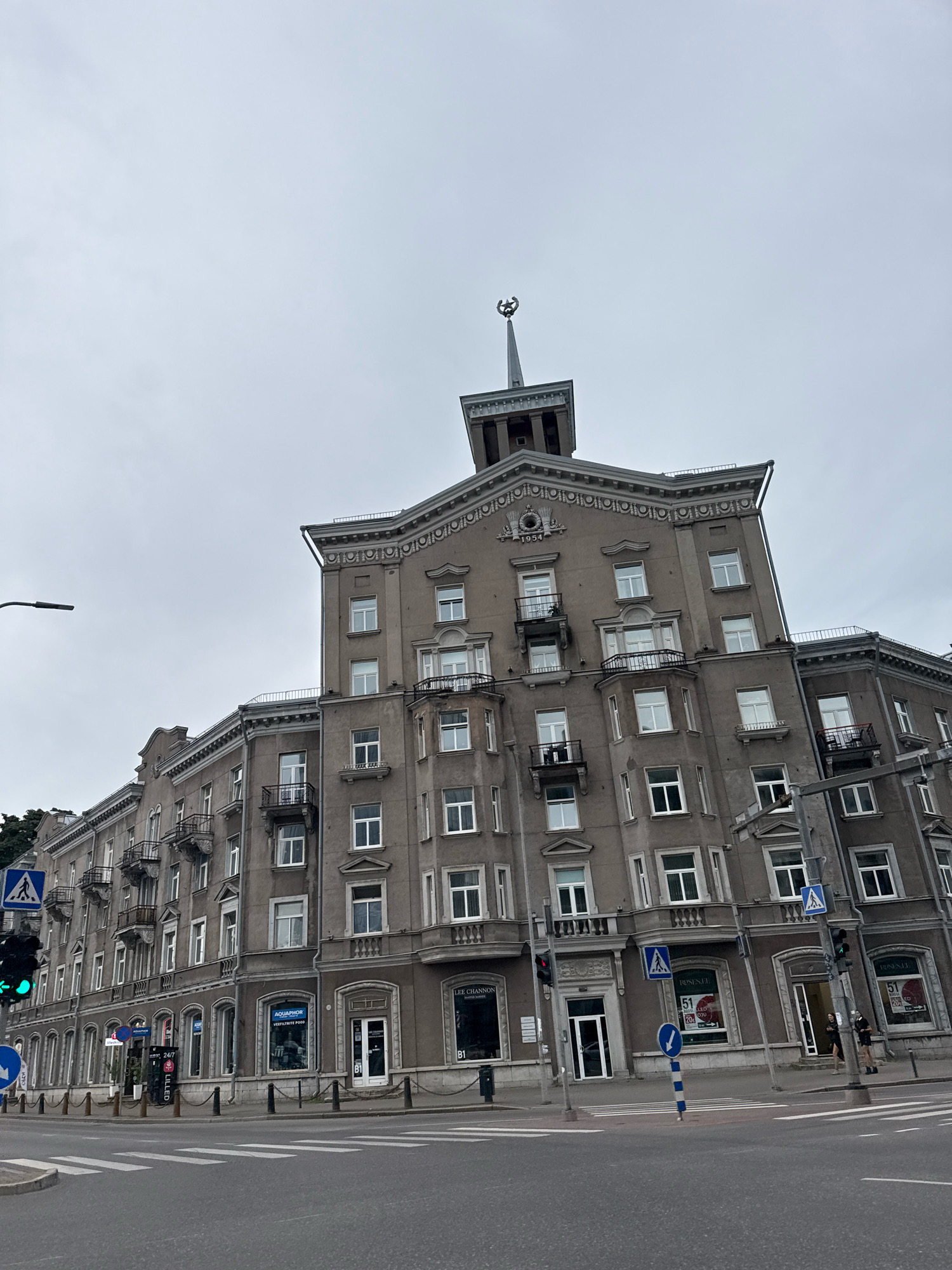 The image shows a street scene in Tallinn, Estonia, featuring a large, multi-story building with an elaborate facade and a distinctive spire topped with an emblem. The building, which appears to be of Soviet-era architecture, has numerous windows, balconies, and decorative elements. A street with a pedestrian crossing is visible in the foreground, along with traffic lights and street signs. A few people are present on the sidewalk in front of the building. The sky is overcast, suggesting a cloudy day. The building's architecture includes classical influences and a prominent central tower, typical of some Stalinist Empire style buildings. The year "1954" is inscribed above a central window, indicating its construction date. Shops are located on the ground floor, with signage for "Aquaphor," "Lee Channon," and "Roses.ee." The overall impression is that of an urban environment with a historical architectural context.