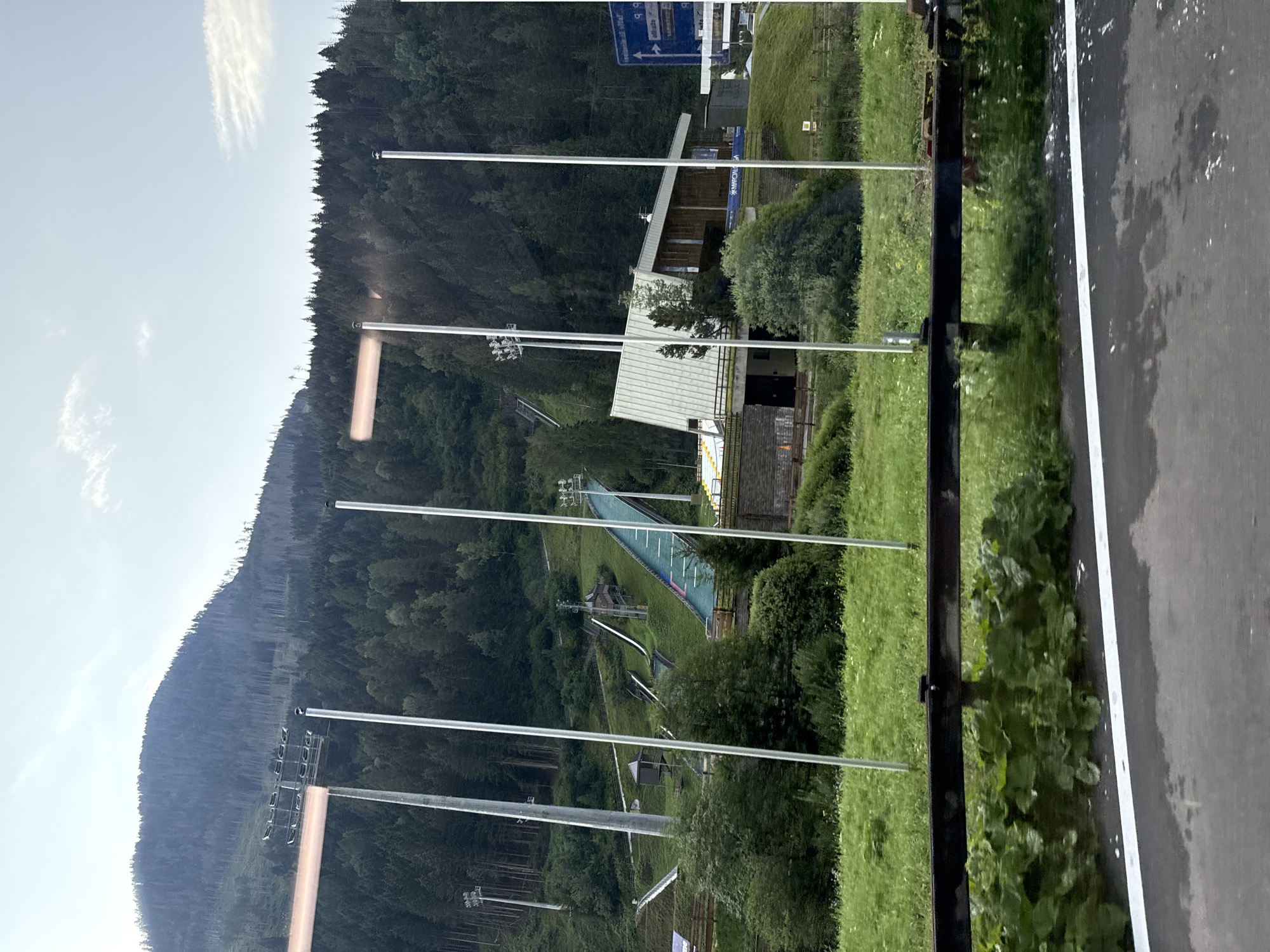 The image captures a ski jumping complex nestled within a mountainous, forested region. The ski jumps, with their steep inclines and green landing surfaces, are visible on the hillside. A building, likely a lodge or administrative center, stands at the base of one of the jumps. Several tall poles with lights are interspersed throughout the scene, suggesting the facility can be used at night. The environment is predominantly green, with lush grass covering the slopes and dense trees forming a backdrop. The sky is partly cloudy, with a clear blue expanse visible in the upper portion of the frame. The image appears to be taken from a moving vehicle, indicated by the blurred foreground and the presence of a guardrail. The ski jumping complex is located in Predazzo, Italy, a town known for its winter sports facilities.