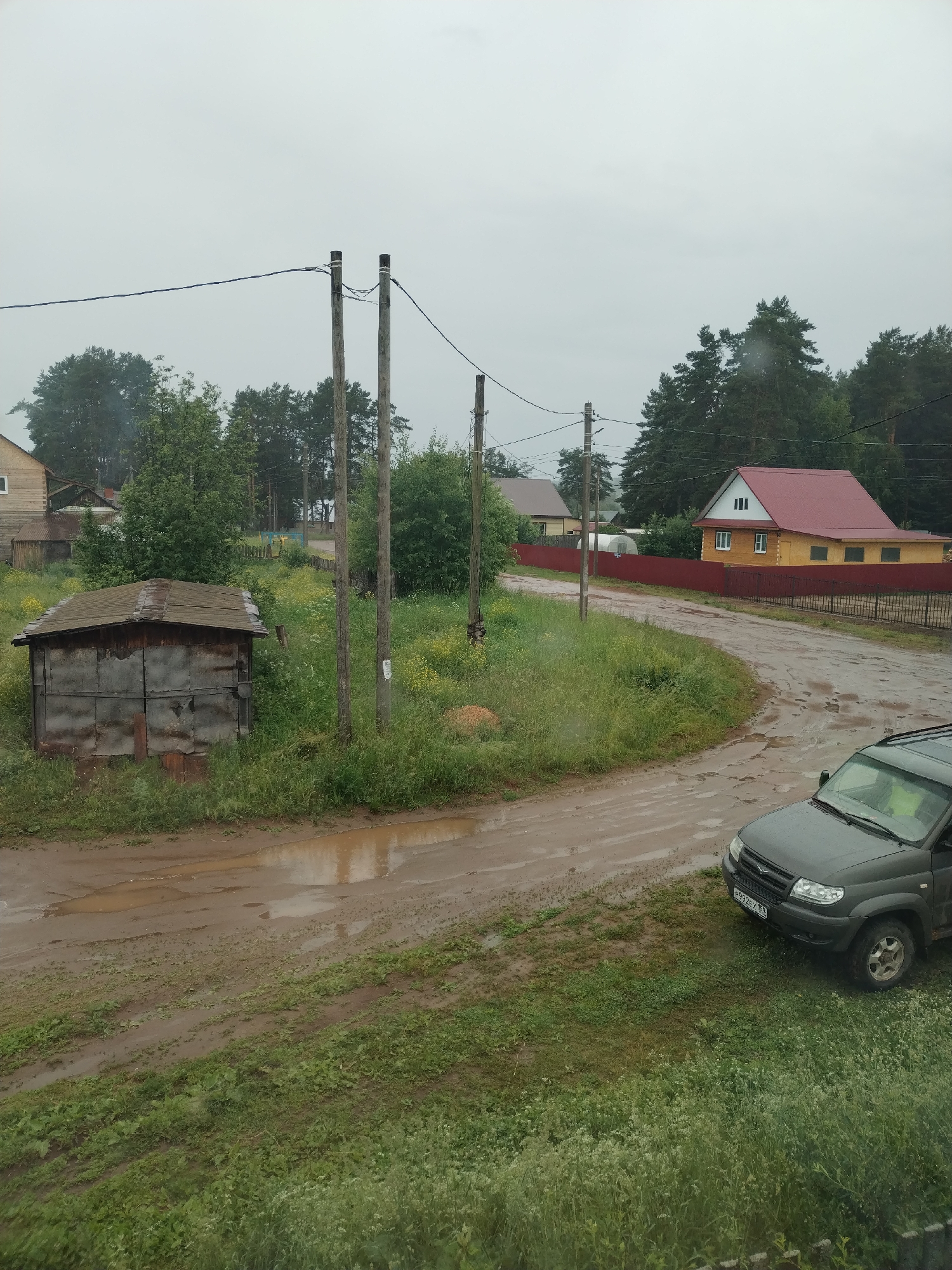 The image depicts a rural scene in Rusalevka, Russia, on a cloudy day. In the foreground, a grey SUV is parked on the side of a muddy, wet road. The road curves to the left, with puddles reflecting the overcast sky. To the left of the road, a small, weathered wooden shed with a metal roof sits amongst tall grass and wildflowers. Further back, a red-fenced property contains a two-story house with a yellow facade and a red roof. In the background, several utility poles with overhead wires are visible, and a dense line of trees, primarily pines, borders the landscape. Another house, lighter in color, is partially visible behind the shed. The overall impression is one of a damp, rural environment, possibly after rainfall.