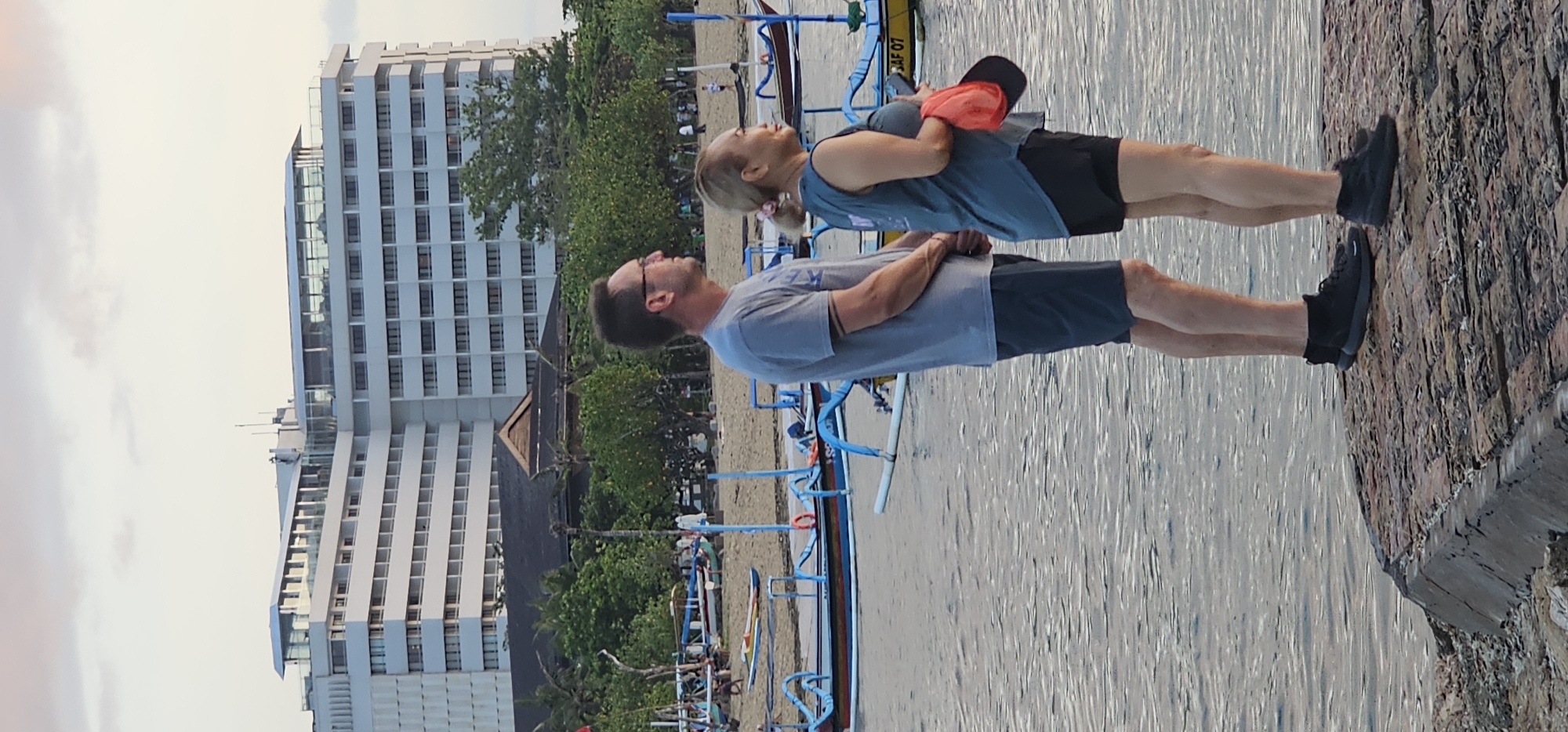 A couple stands on a stone ledge in Denpasar, Indonesia. The man, on the left, wears a gray t-shirt, dark shorts, glasses, and black shoes. The woman, on the right, has blonde hair in a ponytail, a gray tank top, dark shorts, and black shoes. She is also holding an orange object in her left hand. Both individuals are looking upwards.
Behind them is a body of water with blue-colored outrigger boats. Beyond the boats is a sandy beach, green trees, a traditional building, and a tall, modern, light-colored hotel. The sky is a pale orange-gray.