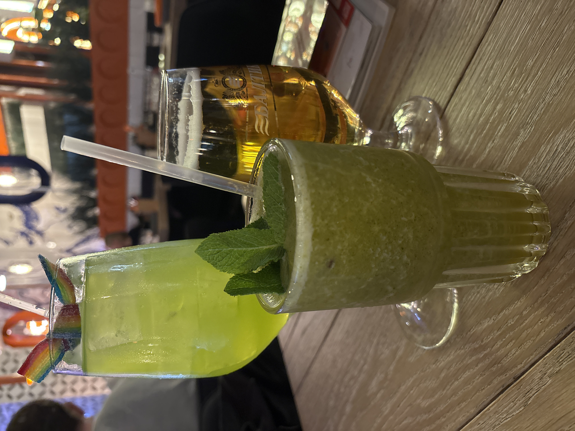 The image displays a close-up, slightly overhead view of three drinks on a light wooden table within an indoor setting, likely a restaurant or bar in Vsevolozhsk, Russia.
In the foreground, two cocktails are visible. On the left, a bright, translucent yellow-green drink in a tall, ridged glass is garnished with a colorful rainbow candy attached to a clear straw. To its right, another green drink, appearing thicker and more textured like a smoothie or slushy, is presented in a similar ridged glass, adorned with fresh mint leaves. Partially obscured behind and to the right of these cocktails is a glass of amber-colored beer, topped with foam. A clear plastic straw extends from the left cocktail, passing in front of the beer glass.
The background is softly blurred, showing warm, artificial lighting elements and indistinct architectural features, indicating an indoor evening or night ambiance. The wooden table features a visible grain pattern. The activity depicted is a casual gathering where drinks are being enjoyed.
A notable detail is the text "БАЛТИКА" (Baltika), written in gold Cyrillic script, clearly visible on the side of the beer glass. Baltika is a well-known Russian brewing company, which aligns with the provided location context of Vsevolozhsk, Russia. No specific people are in focus, but their presence is implied.