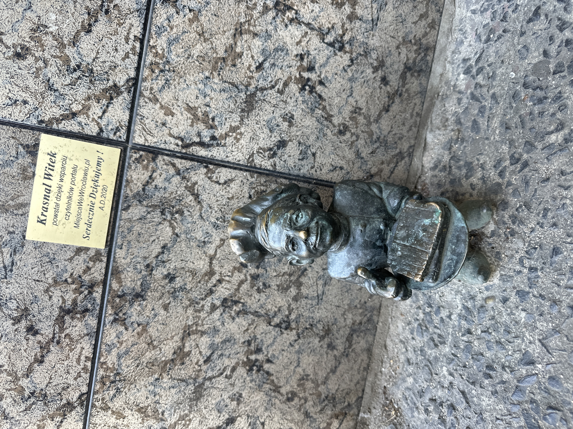 The image depicts a bronze statue of a Wrocław dwarf, located on a cobblestone surface, next to a plaque. The dwarf, wearing a chef's hat, holds a plate with a book on it. The plaque, with text in Polish, is attached to the pavement with a thin metal bar. The surface around the statue and plaque is a mix of stone tile and cobblestone. The lighting suggests an outdoor setting.