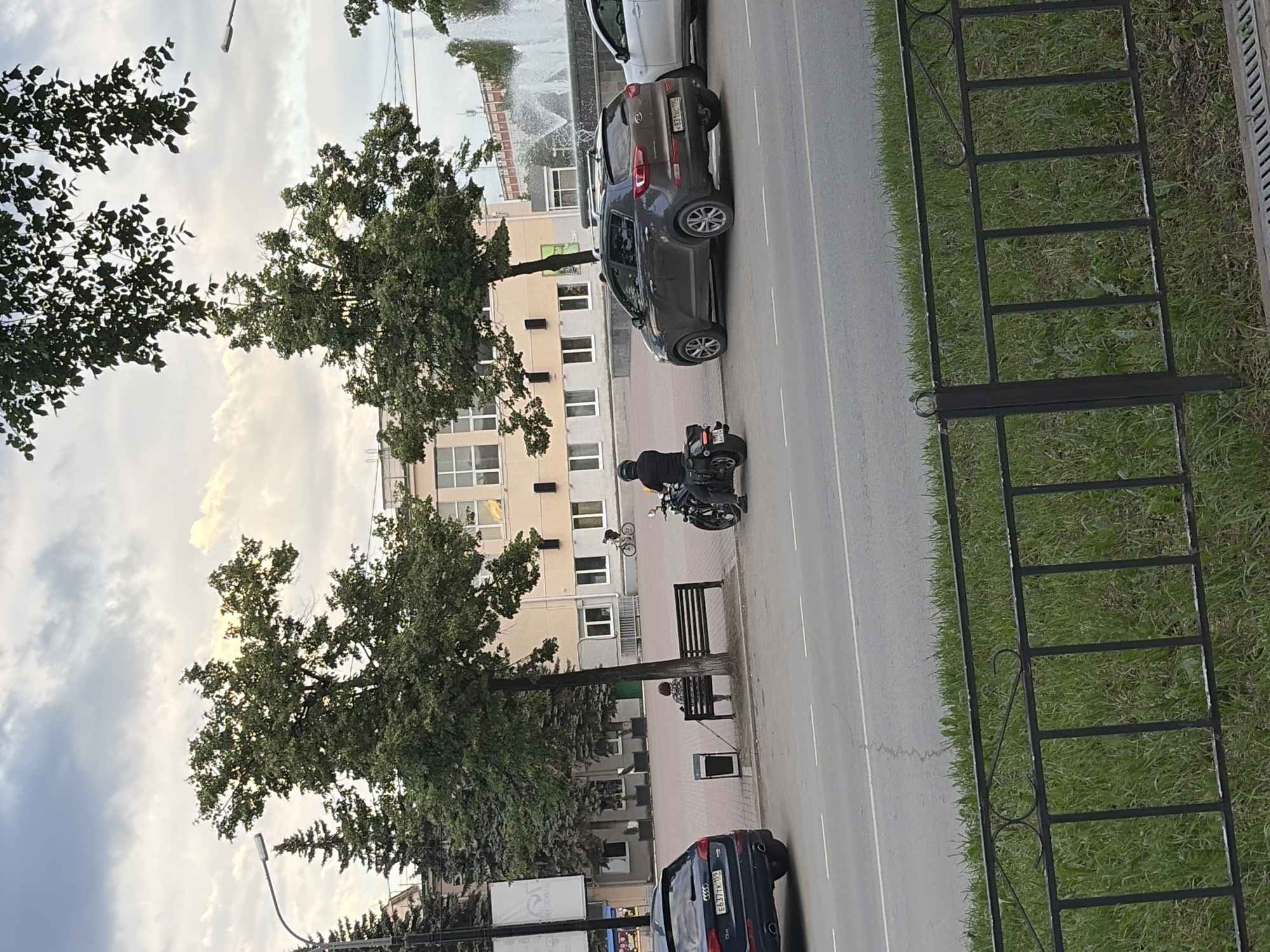 The image depicts a street scene in Chaikovsky, Russia, likely in the late afternoon or early evening, given the soft lighting and overcast sky. The orientation of the image is rotated 90 degrees counter-clockwise.
The foreground features a dark-colored sedan parked on the right side of the road, with its license plate clearly visible: E837TK 68. To the left of the sedan, a black metal fence with decorative elements separates the sidewalk from a grassy area. Further to the left, a sidewalk runs alongside a road, with a lone tree providing shade. A woman is seated on a dark-colored bench near the tree, facing away from the camera. Beside the bench is a dark trash receptacle.
Further down the sidewalk, a motorcycle is parked, and a person wearing a dark helmet and jacket is seated on it, looking towards the right. In the background, a multi-story building with light-colored walls and numerous windows stands. Several trees with lush green foliage are scattered throughout the scene. The sky is a mix of clouds and patches of lighter sky. A fountain is visible in the far background. The overall impression is a quiet, urban street with elements of nature interspersed.