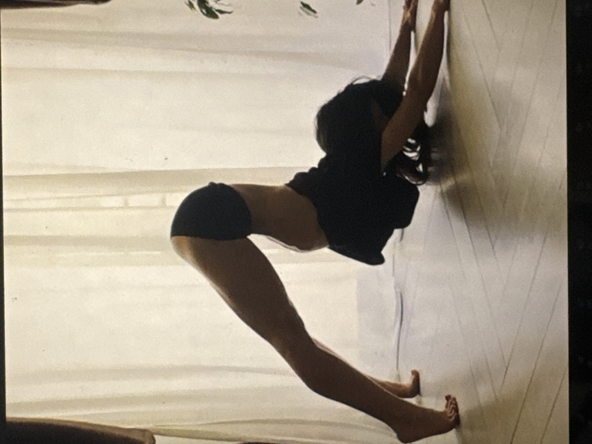 A person is performing a yoga pose, specifically a variation of downward-facing dog, on a light-colored wooden floor in what appears to be an indoor setting with sheer curtains in the background. The person is wearing dark athletic shorts and a dark top. Their body is in an inverted V-shape, with hands and feet on the floor and hips raised. The lighting suggests natural light coming from behind the person, creating a silhouette effect. There is a potted plant visible on the right side of the frame. The image quality is moderate with some graininess. The specific location of Kuta Selatan, Indonesia, cannot be determined from the visual information provided in the image.