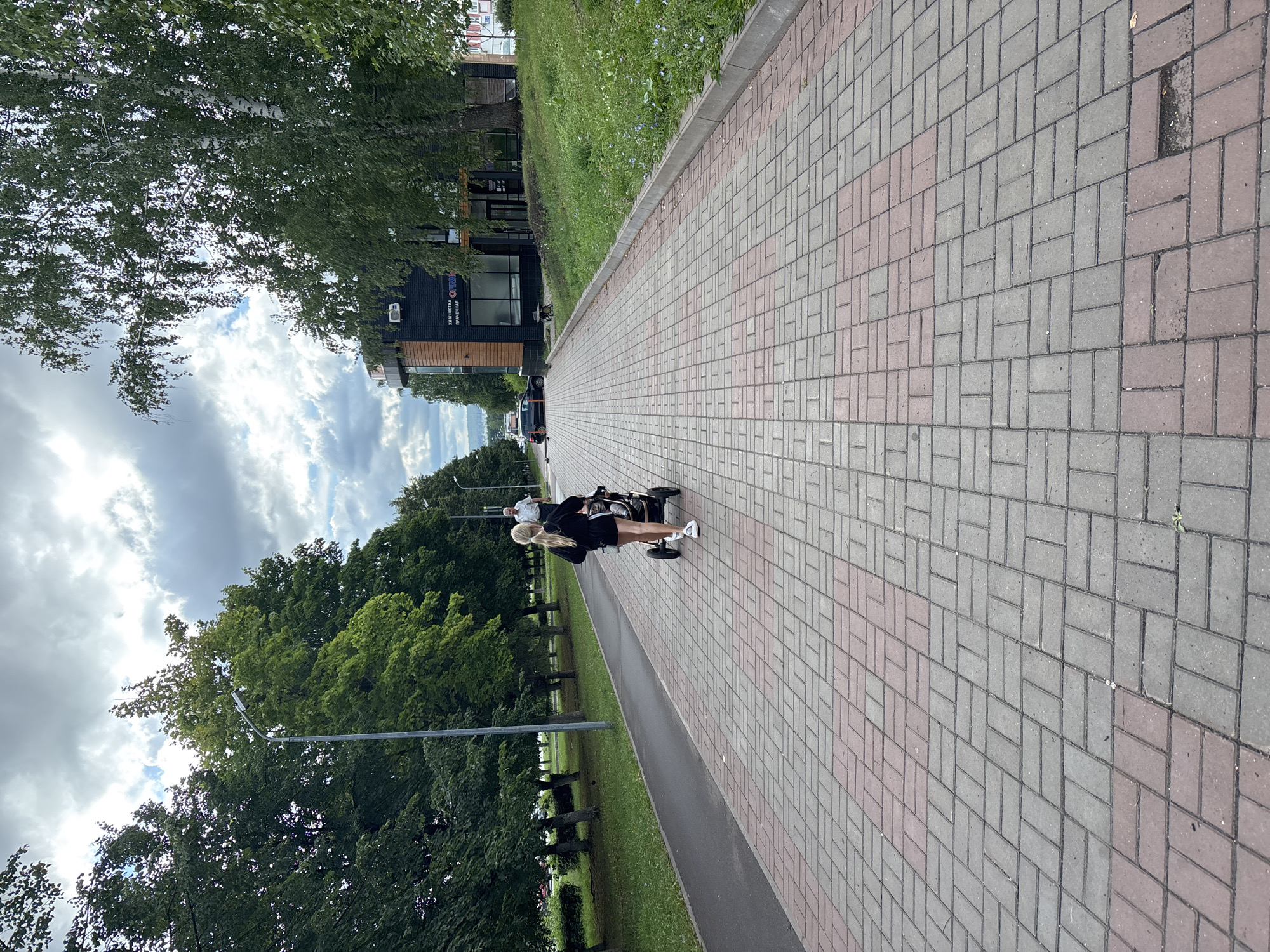 A young woman with blonde hair pulled back in a ponytail is pushing a stroller along a paved walkway. She is wearing a black top and shorts, and white sneakers. The walkway is made of red and grey pavers, arranged in a herringbone pattern in some areas and a basketweave pattern in others. To the left of the walkway is a grassy area with trees. To the right of the walkway is a building with dark siding and a wooden accent wall, and a green grassy slope with scattered wildflowers. A man in a light-colored shirt and dark pants is walking in the distance on the same walkway, ahead of the woman. The sky is cloudy. The building appears to be a commercial establishment with Cyrillic lettering that reads "ХИМИЧИСТКА ПРАЧЕЧНАЯ" which translates to "Dry Cleaning Laundry". The scene is likely in Naberezhnye Chelny, Russia, given the building's signage.