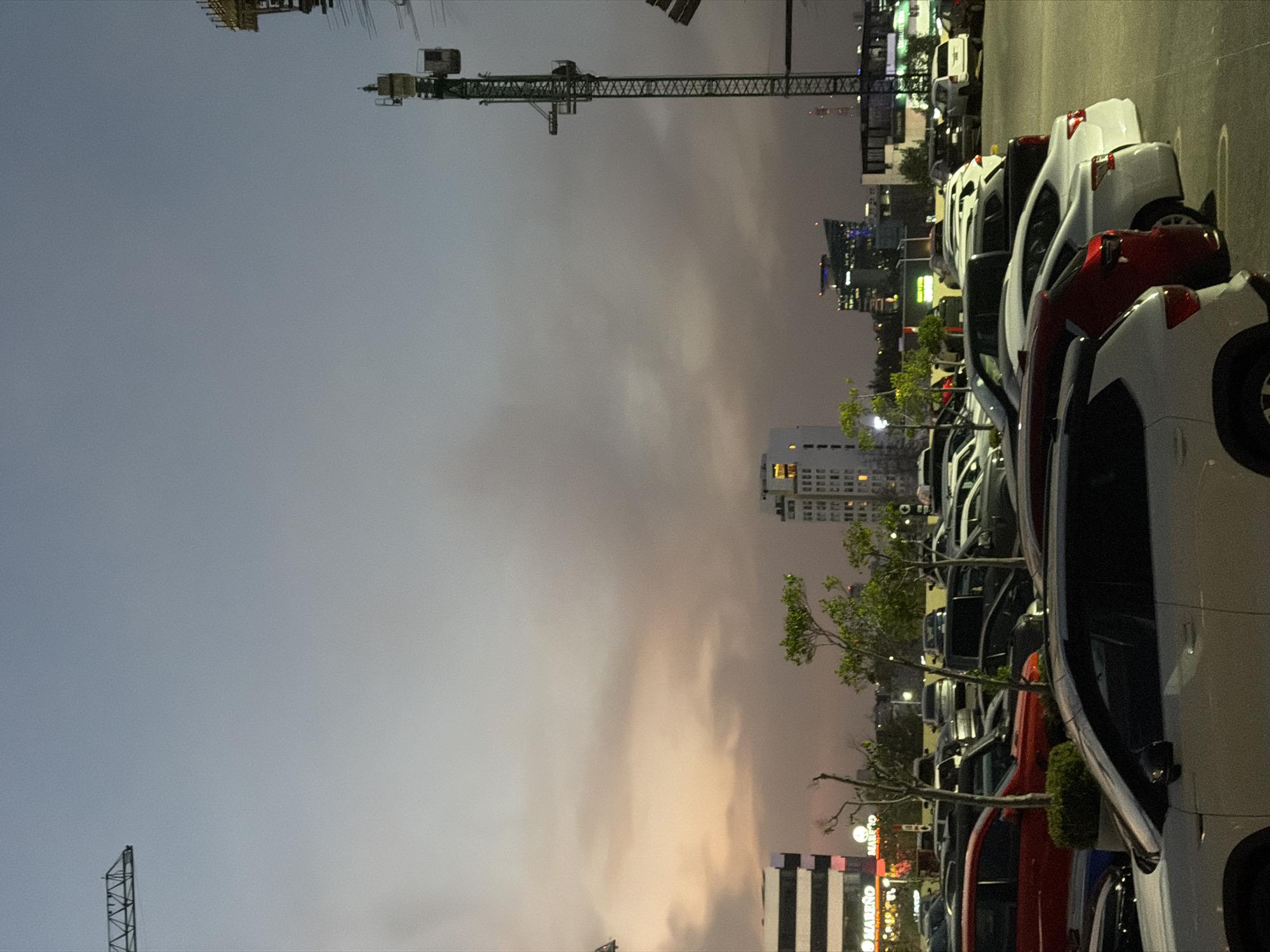 The image shows a parking lot in the city of Puebla, Mexico, at dusk. The lot is filled with parked vehicles, primarily cars and trucks, lined up in rows. A construction crane is visible in the background, along with several buildings, including a high-rise with lit windows. The sky is a mix of gray and orange, suggesting either sunset or a heavy overcast. Trees are interspersed between the parked cars, adding a touch of greenery to the urban scene.