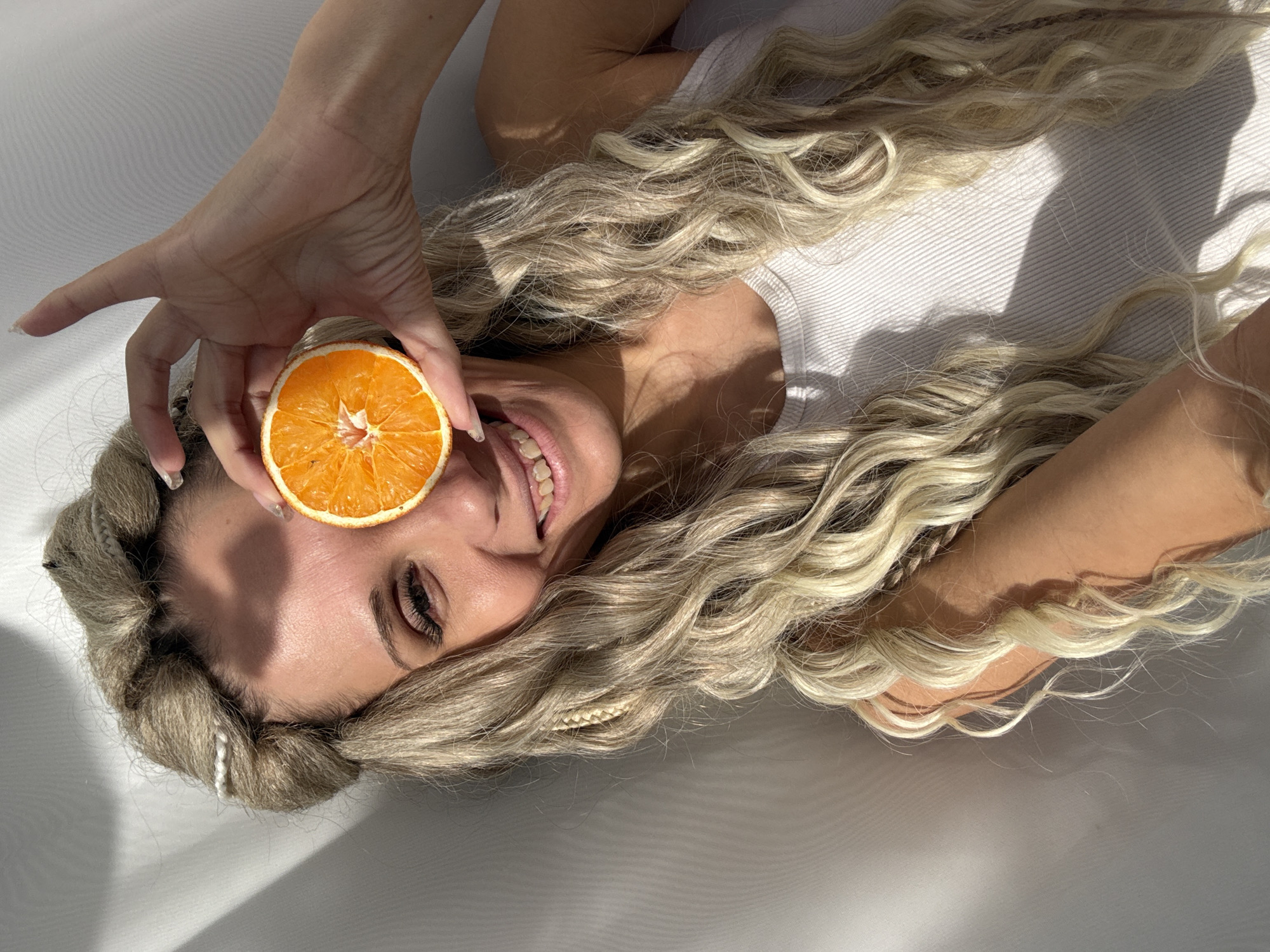 A woman with long, wavy blonde hair, partly styled into braids, lies on her back on a white, textured surface. Her head is angled slightly upwards, revealing a broad smile with visible teeth. She holds a half-slice of a dried orange over her right eye with her left hand, obscuring it. Her left eye is open and looking towards the camera. Strong, defined shadows are cast across her face and the white background, suggesting direct daylight or a powerful artificial light source, indicating it is daytime. She is wearing a white ribbed tank top or similar garment. The overall expression on her face is cheerful and playful. There is no visible text in the image.