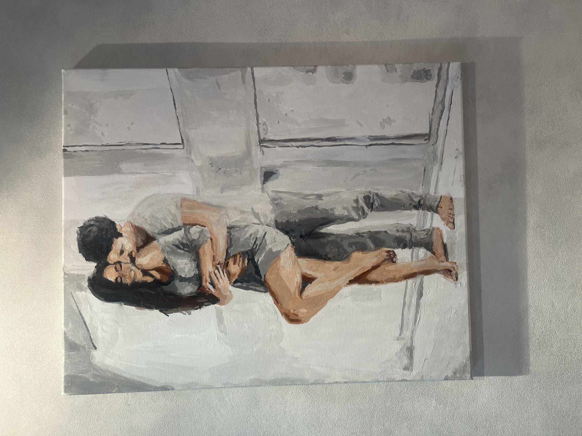 An oil painting on canvas depicts a couple embracing. The woman, with long dark hair, is leaning back into the man, her legs crossed. She is wearing a grey top and dark shorts. The man, with dark curly hair, is wearing a grey t-shirt and grey pants. They are standing indoors, in what appears to be a minimalist room with white and grey walls. The lighting is soft, casting a shadow on the right side of the painting. The painting is hung on a textured wall. There is no indication of the location being in the city of Rybnitsa, Moldova.