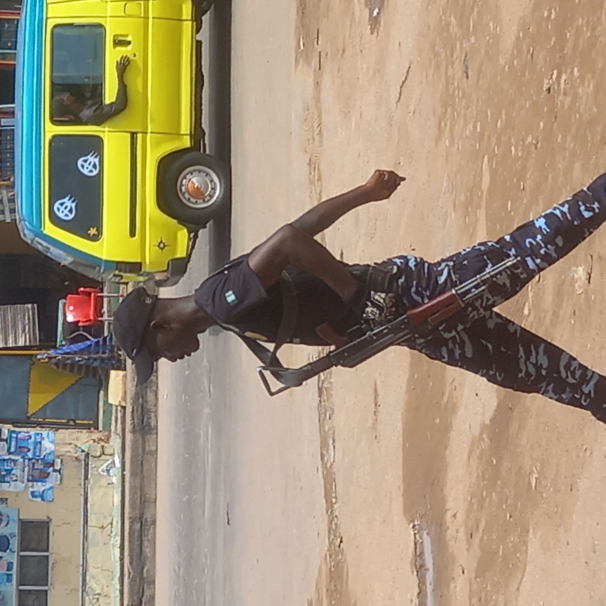 A man in camouflage pants and a black shirt, wearing a cap, is walking and carrying a rifle in Dadi Riba, Nigeria. He appears to be crossing a street. A yellow van with a blue top is parked on the side of the street, with a person visible inside. The ground is paved. A building and other objects are visible in the background.