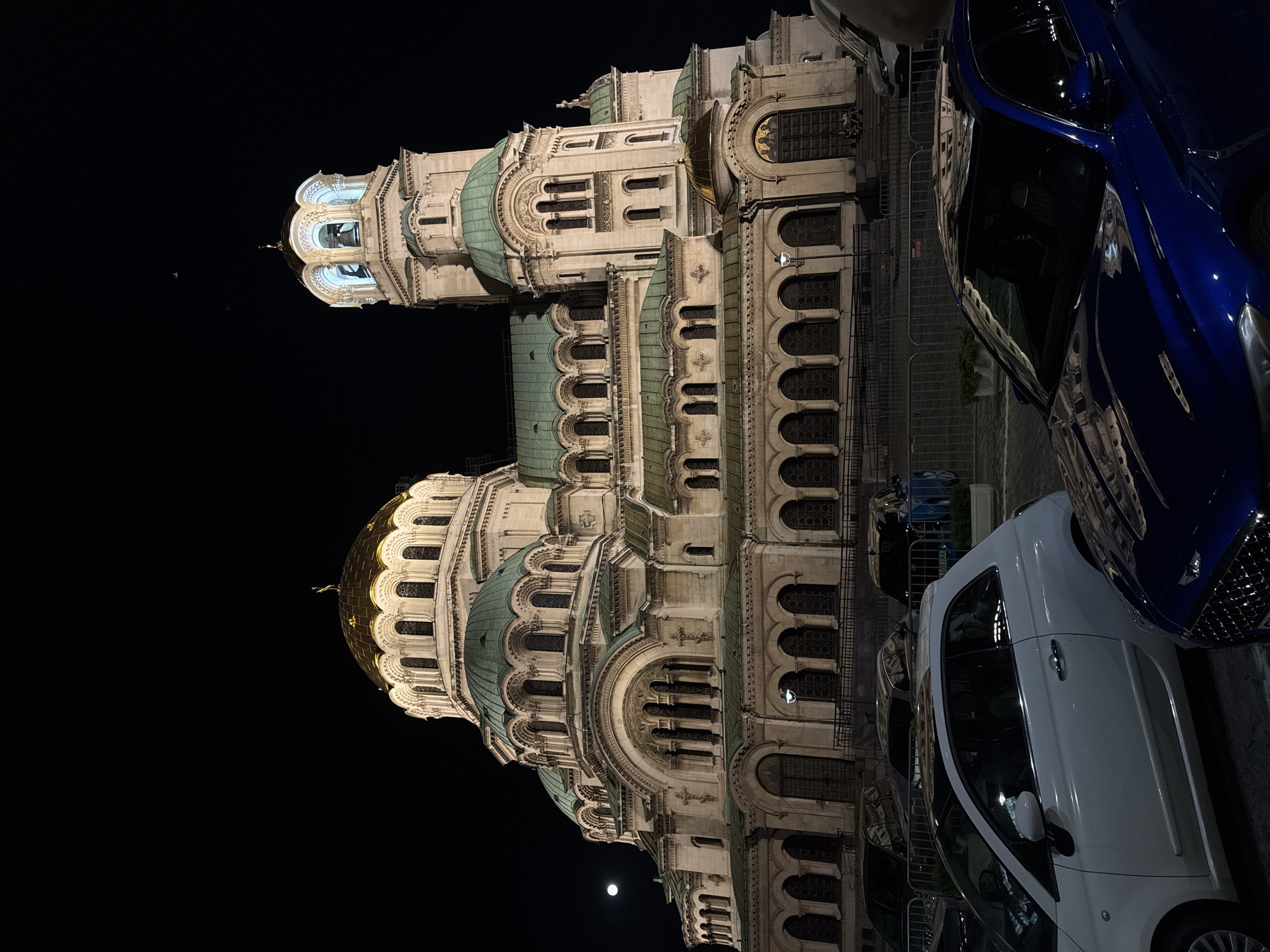 The image depicts the Alexander Nevsky Cathedral in Sofia, Bulgaria, at night. The cathedral, a prominent landmark, is illuminated, showcasing its intricate architecture with multiple domes, including a large golden central dome and a bell tower. The sky is dark, with a full moon visible on the left side of the image. In the foreground, several cars are parked. A white Fiat 500 is on the left, and a dark blue sedan is in the center-right. A metal fence separates the parking area from the cathedral's entrance, where a few individuals are seated on the steps. The lighting creates a dramatic contrast between the illuminated building and the dark surroundings, highlighting the cathedral's grandeur.