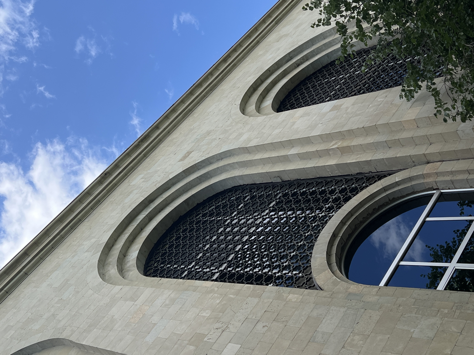 An upward-angled shot captures a section of a light-colored stone building, likely in Yerevan, Armenia, beneath a bright blue sky with scattered white clouds. The facade features several distinctive architectural elements: two large, recessed arched windows covered with intricate dark metal grilles displaying an ornamental, repeating geometric pattern, and a prominent circular window with a simple cross-shaped frame. This circular window reflects the blue sky and clouds, along with some green foliage from a nearby tree.
The building's surface is constructed from uniform, light beige or cream-colored stone blocks, possibly tufa or travertine, characteristic of Armenian architecture. Curved stone ledges and layered relief work frame the arched windows, adding depth and texture to the facade. Green leaves from a tree are visible in the upper right corner, partially obscuring the top of the building and casting reflections onto the circular window. The overall impression is one of architectural grandeur and detail, bathed in bright daylight under a clear, sunny sky. No human activity or visible text is present in the image.