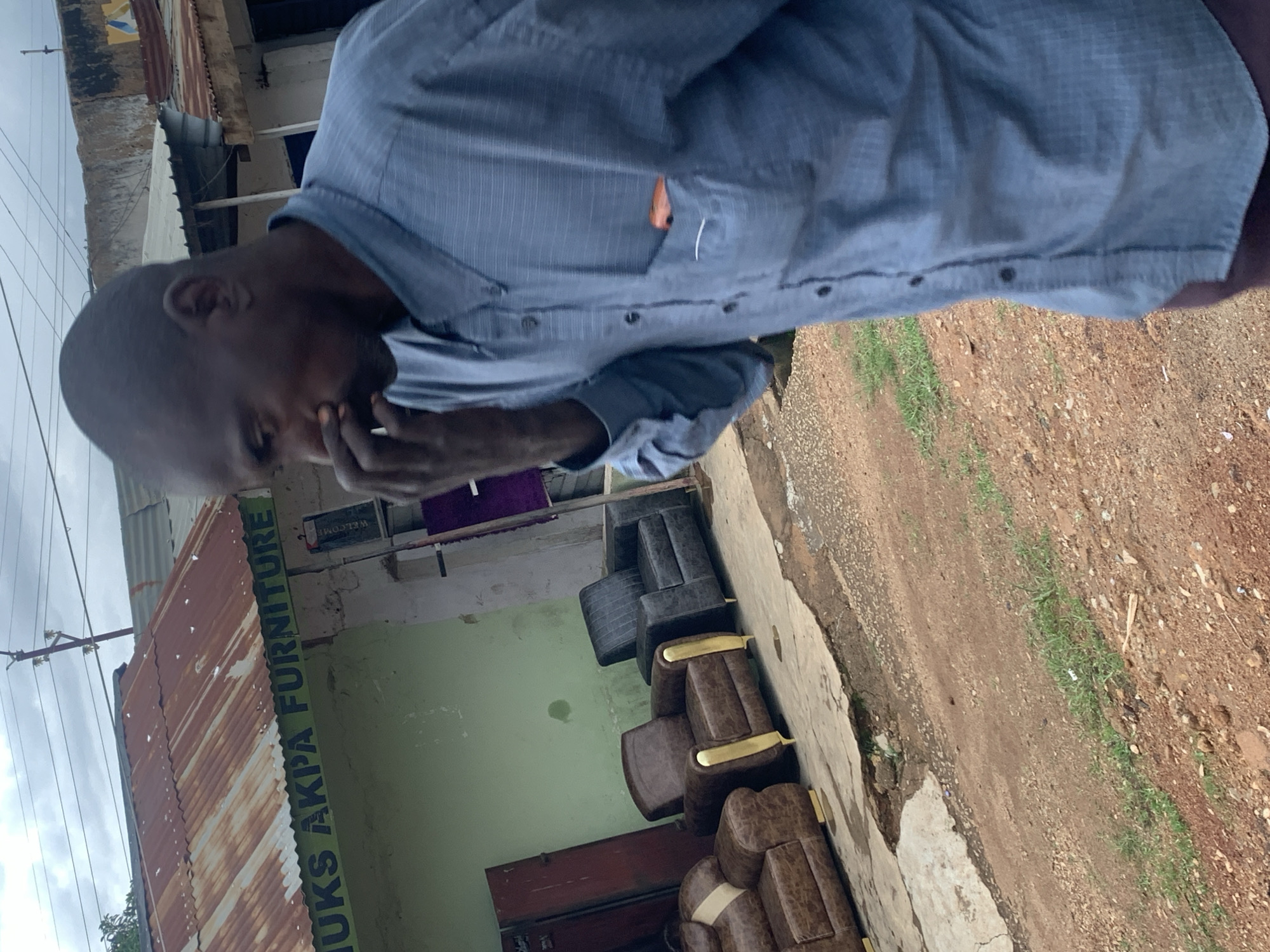 The image shows a man in a blue pinstripe shirt with his hand to his face, suggesting contemplation or discomfort. He appears to be standing outside a building with a corrugated iron roof. In the background, there is a store with the visible text "JUKKS AKIPA FURNITURE" painted vertically on a light green wall. The store appears to be selling furniture, as several chairs and sofas are visible outside the entrance, some with gold accents. The ground is a mix of dirt and grass, and the sky is overcast, suggesting it might be a cloudy day. The scene appears to be in a commercial area in Jos, Nigeria. The overall impression is one of an everyday scene in a marketplace or commercial street.