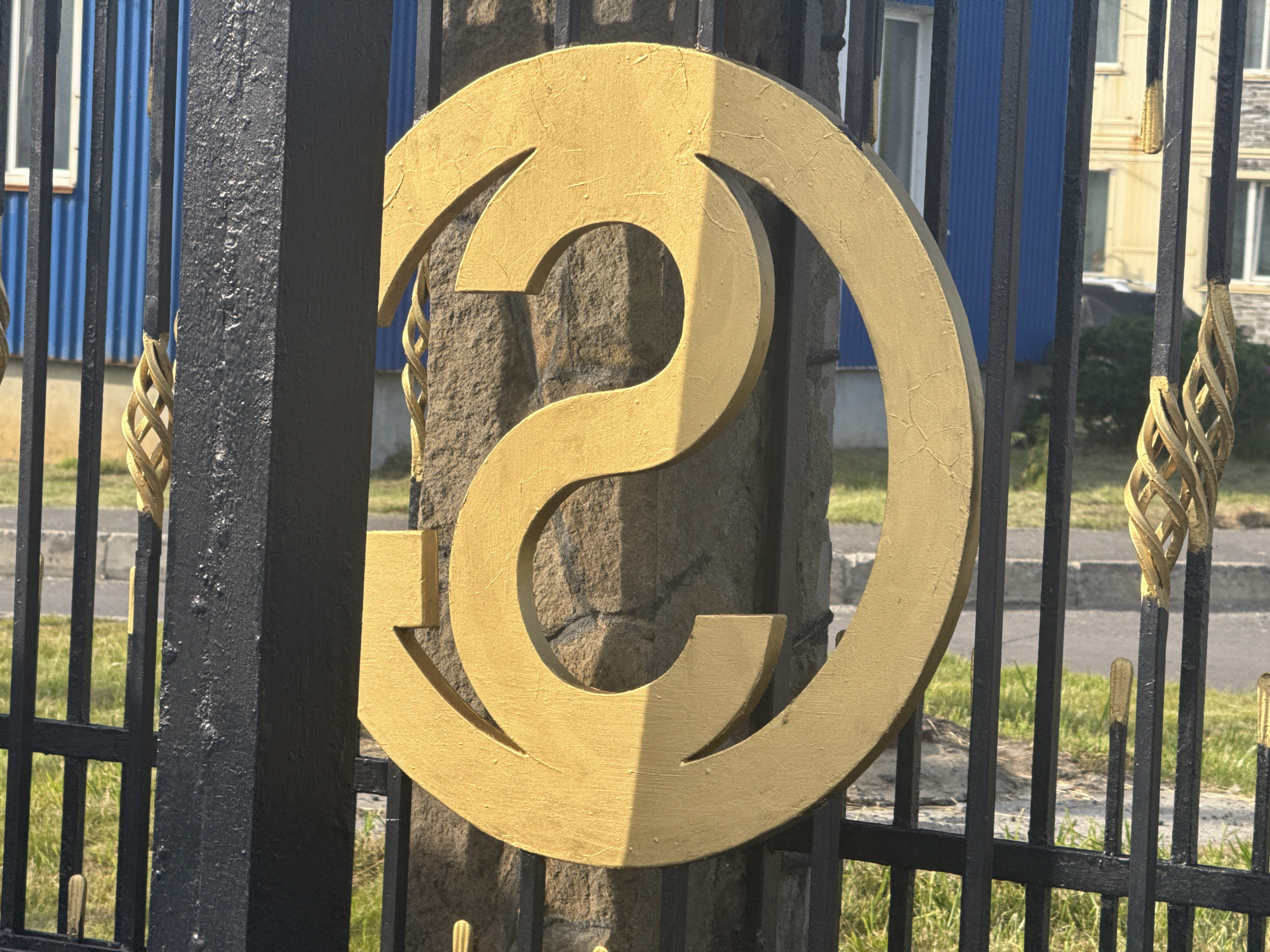 A close-up, eye-level shot shows a portion of a black metal fence with ornate gold accents, partially obscuring a large, golden, stylized letter or symbol. The symbol appears to be a thick, curved band with a cut-out section, resembling a stylized "2" or an abstract "S" shape. It is affixed to a rough, stone-like pillar behind the fence. The fence itself has vertical bars and some decorative twisted gold elements. In the background, parts of buildings are visible, with blue and yellow siding and white-framed windows. The environment appears to be outdoors, with green grass in the foreground and what looks like a sidewalk or road further back. The lighting suggests it is daytime, with shadows cast by the symbol and fence onto the pillar and surrounding area. This scene is located in the city of Kuril'sk, Russia.