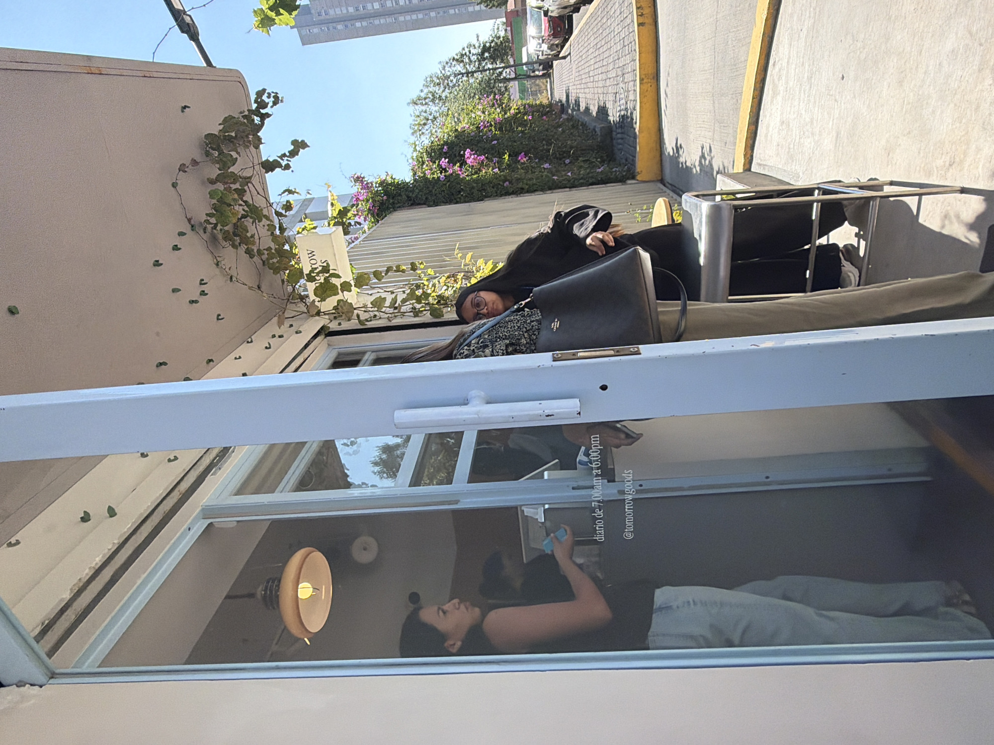 The image depicts a person sitting at a table outside a cafe, reflected in the cafe's window. The cafe appears to be named "Tomorrow Goods," judging from the text "@tomorrowgoods" visible on the window. The reflection shows another person inside the cafe, appearing to be using a phone. The location is likely Puebla, Mexico, based on the text "diario de 7:00am a 6:00pm" on the window, which is in Spanish. The environment includes a sidewalk, street, and building in the background. There are also plants and vines growing near the cafe.