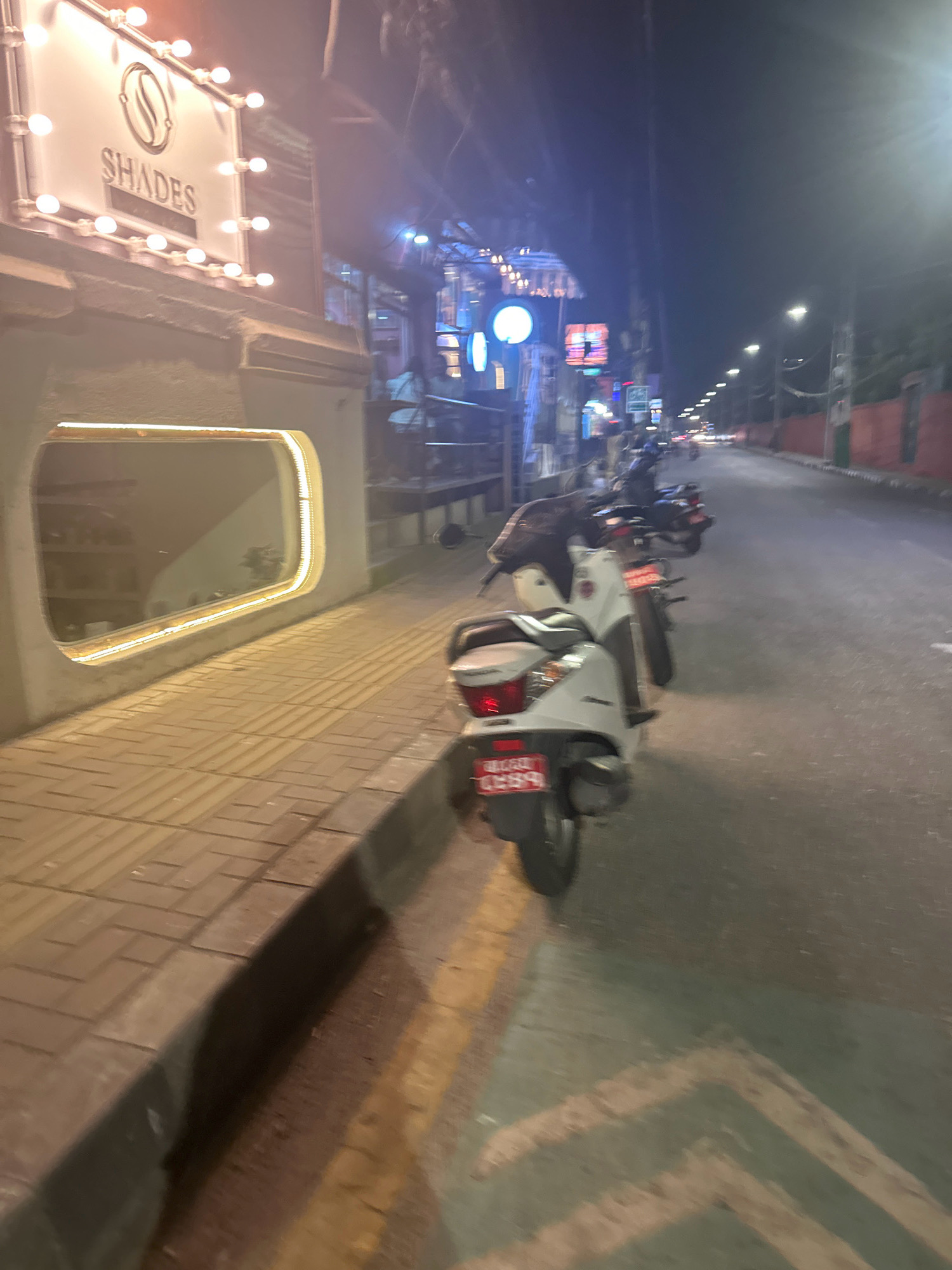 At night in Lalitpur, Nepal, a street scene unfolds with a focus on a building facade and a row of parked motorbikes. The building features a prominent illuminated sign reading "SHADES" above a distinctive circular logo. The facade also has a large, irregularly shaped window with an internal amber light. Adjacent to the building is a sidewalk paved with rectangular tiles, demarcated by a raised edge.
A line of motorbikes is parked on the side of a paved road that extends into the distance. The closest motorbike is white and positioned on the sidewalk, partially obscuring the tiled pathway. Its rear license plate is visible. Further down the road, additional motorbikes are parked in a line, with blurred headlights of approaching vehicles in the background, suggesting ongoing traffic. Streetlights illuminate the scene. The overall atmosphere is dimly lit, characteristic of an urban environment at night.