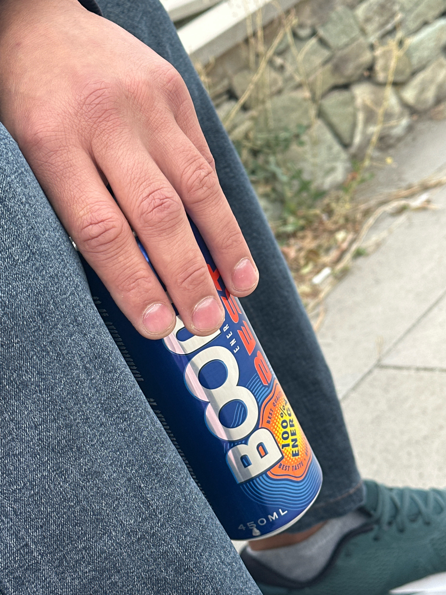 The image shows a close-up view of a person's hand resting on a blue energy drink can. The person is wearing blue jeans. The background appears to be an outdoor setting with stones and dry grass. The energy drink can has the brand name "BOOST" in large white letters. There is also text on the can indicating "BEST QUALITY" and "100% ENERGY," along with "BEST TASTE" and "450ML." The lighting suggests it is daytime.
