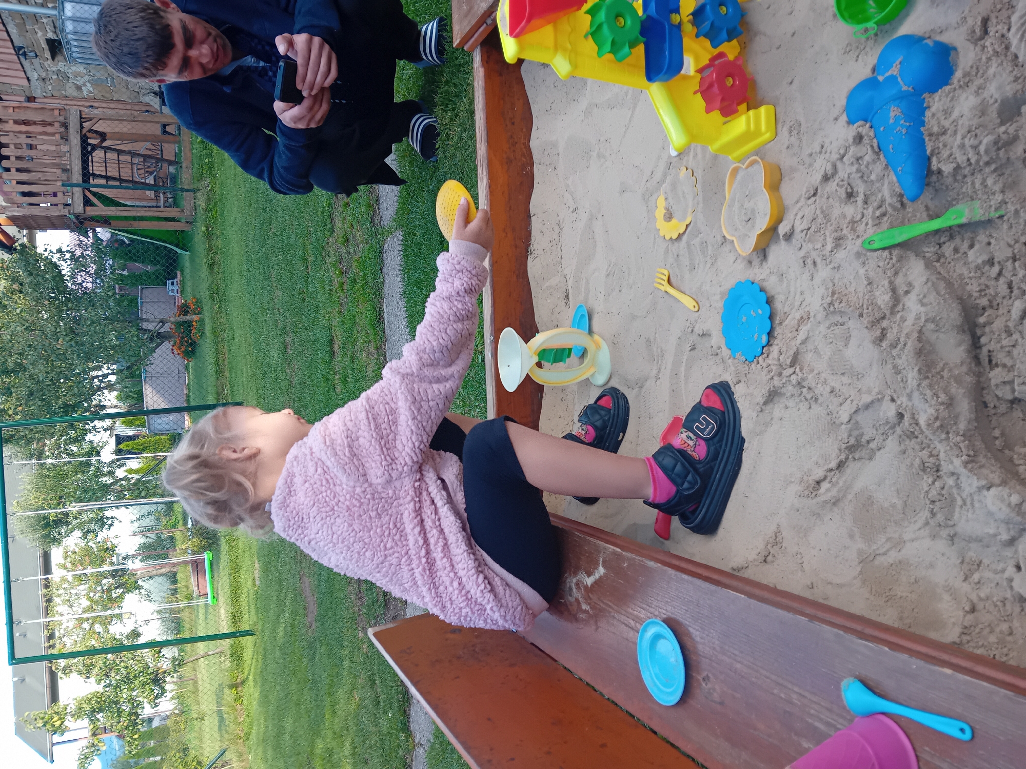 A young girl with blonde hair is sitting on a wooden bench next to a sandbox. She is wearing a pink fluffy sweater, black shorts, and black sandals with pink socks. She is holding a yellow sand toy. In the sandbox, there are various colorful plastic toys, including a yellow sandcastle mold, a blue ice cream cone mold, and a green rake. A man is visible in the background, looking at his phone. The scene appears to be outdoors, with a grassy yard and trees in the background. The lighting suggests it is daytime. There is no visible text in the image. The location context is Zuberec, Slovakia.