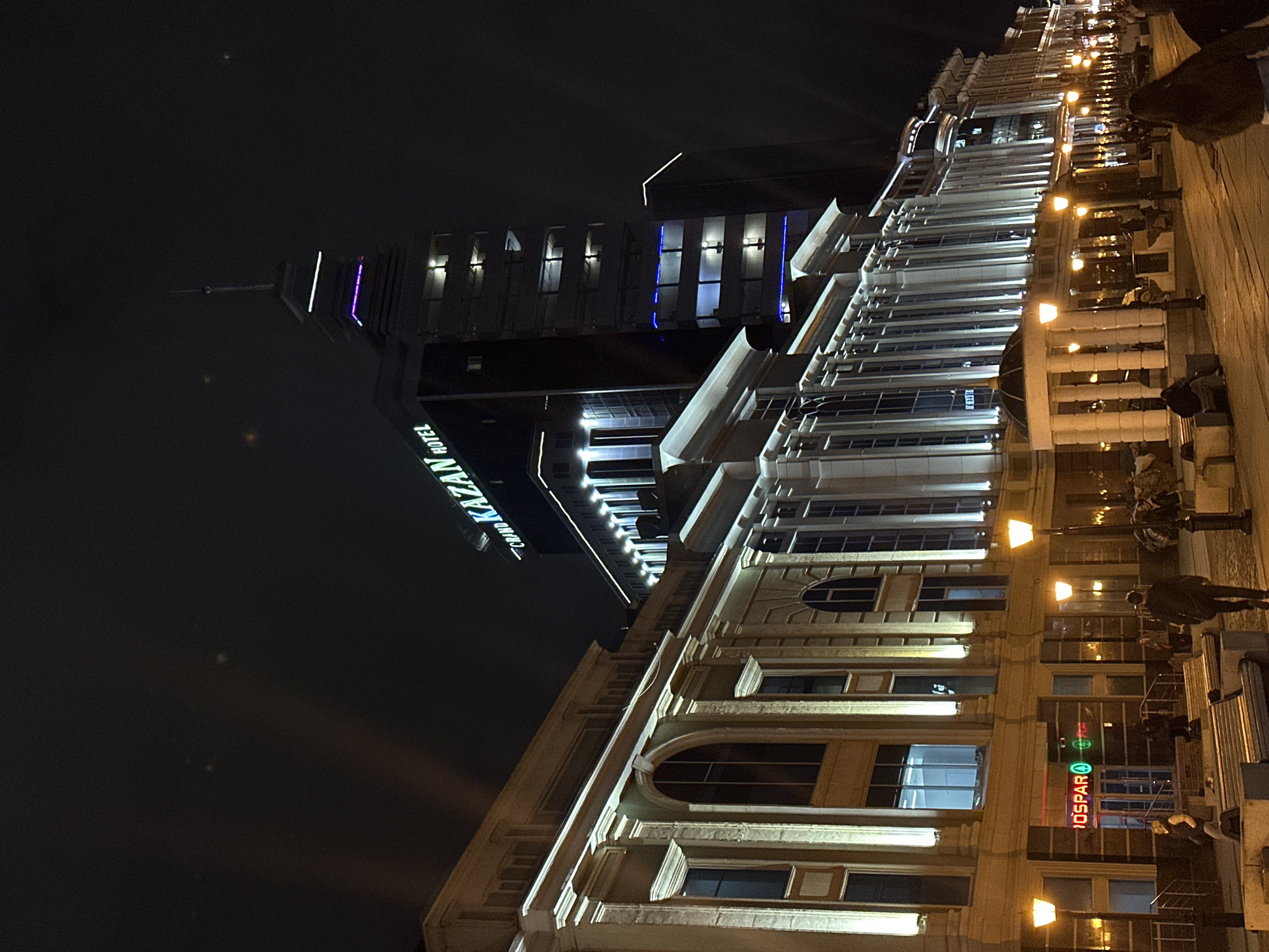 The image, taken at night in Kazan, Russia, shows two distinct buildings. On the left is the "Grand Kazan Hotel," identified by illuminated signage. The other building, adjacent and slightly right, has an older architectural style with multiple stories, ornate window designs, and is illuminated by street lamps. People are seen on the street, with some possibly seated outside the building. A Spar store is indicated by signage. The overall scene depicts an urban environment at night.