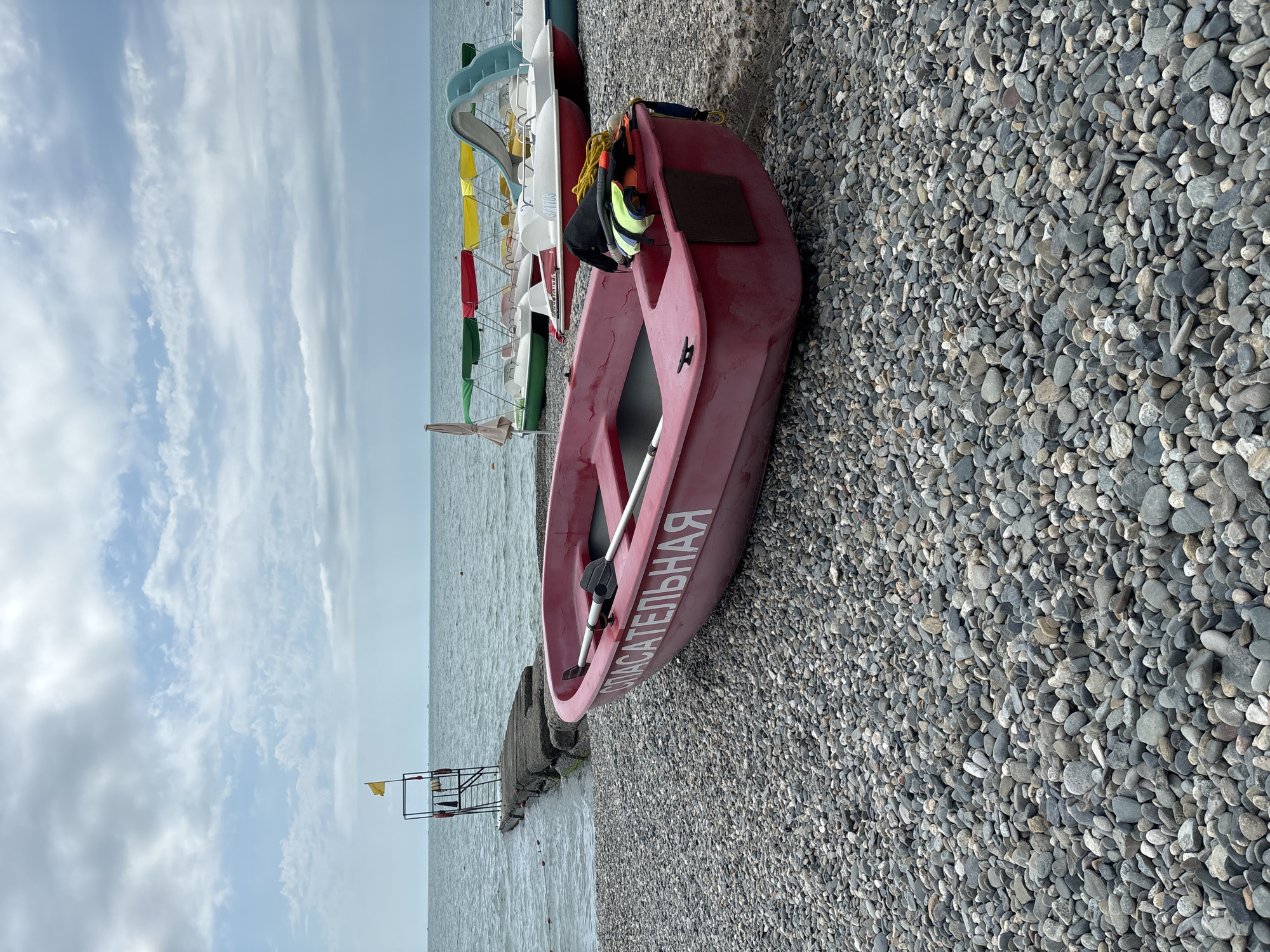 This media file depicts a pebble beach in Sochi, Russia, under a cloudy sky. The primary subject is a red rescue boat resting on the shore, its hull bearing the white Cyrillic text "СПАСАТЕЛЬНАЯ" (Spasatel'naya), meaning "rescue" or "life-saving." A life vest and a helmet are visible inside the boat.
In the background, a row of colorful rental pedal boats with canopies lines the beach, suggesting a recreational area. To the left, a simple metal structure, possibly a lifeguard station or a pier support, is partially submerged in the gentle waves. A yellow flag flies from this structure. The sea is calm, with a hazy horizon. The overall scene conveys a sense of readiness for water activities or a quiet moment before or after them. The time of day appears to be daytime, with diffused light due to the cloud cover.
