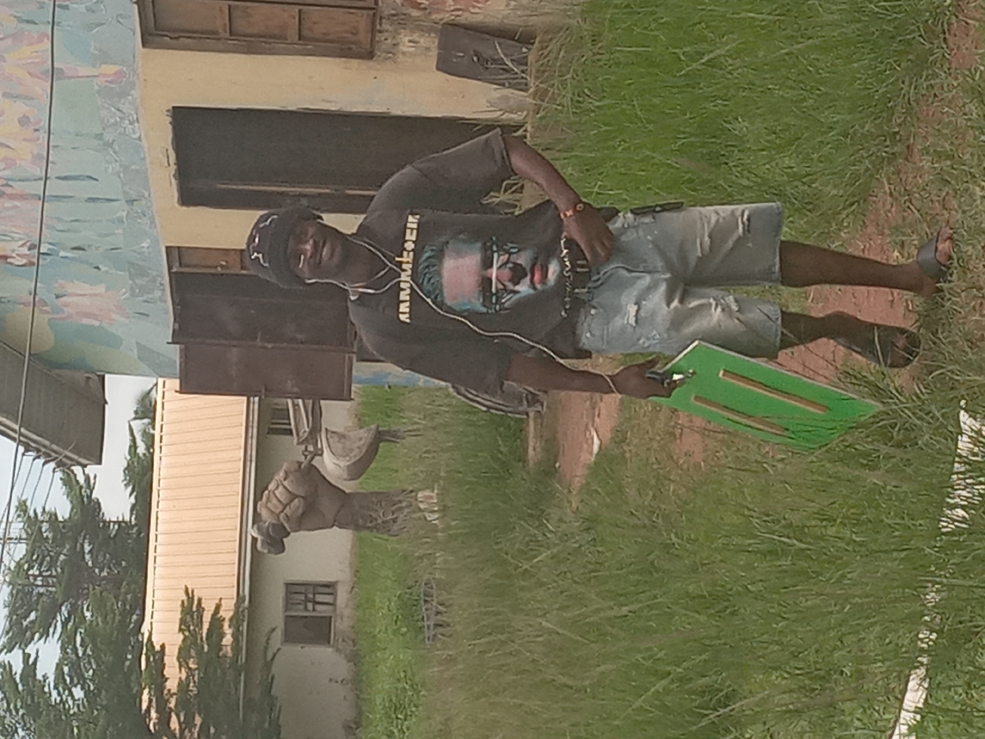 The image depicts a person standing outdoors on a patch of grass. The person is wearing a dark t-shirt with a graphic on it, light-colored denim shorts, a hat, and sandals. They are also wearing headphones. The person is holding a green object, possibly a board or sign. The background includes buildings and vegetation. Based on the provided information, the location is Achara Ndiagu Omegio, Nigeria. A concrete hand is visible in the background.