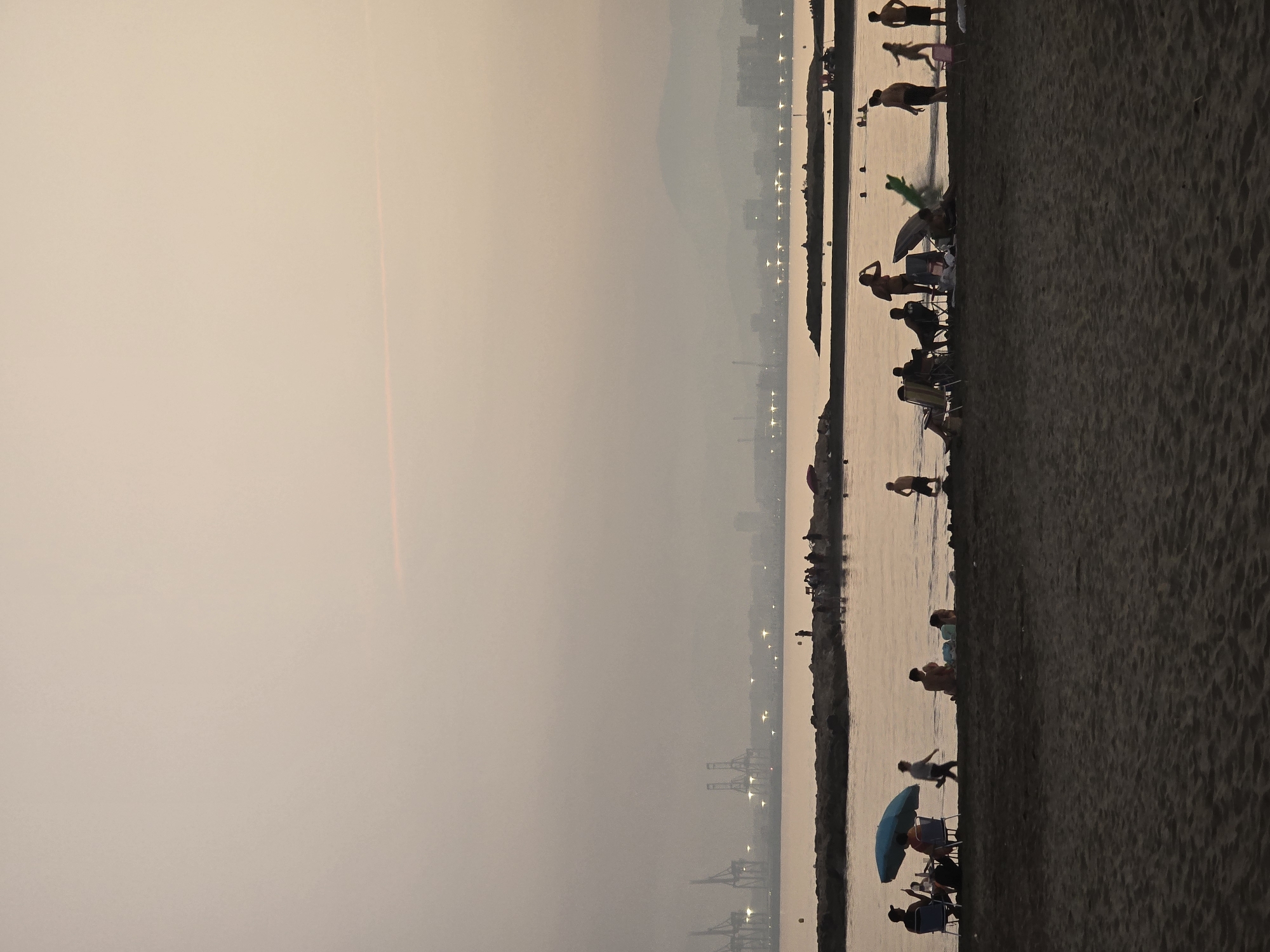 The image captures a serene sunset scene on the beach in the city of Málaga, Spain. Silhouettes of people are scattered across the sandy shore and in the shallow water, suggesting a leisurely evening by the sea. On the beach, individuals are seen sitting on chairs, some under a blue umbrella, while others recline or walk. Children are present, with one appearing to be playing in the water.
In the background, a stone jetty juts out into the calm water, with more figures visible on it, some standing and others sitting. Beyond the jetty, the vast expanse of the Mediterranean Sea stretches towards a hazy horizon. A cityscape with buildings and lights is faintly discernible in the distance, partially obscured by the evening mist. Prominent in the far background are industrial cranes, indicating the presence of a port or industrial area near the city. The overall atmosphere is peaceful, with the soft, diffused light of the setting sun casting a warm glow over the scene.